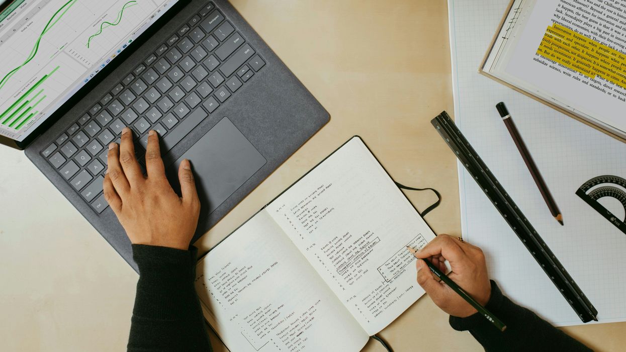 A hand types on a laptop while another takes notes in a notebook.