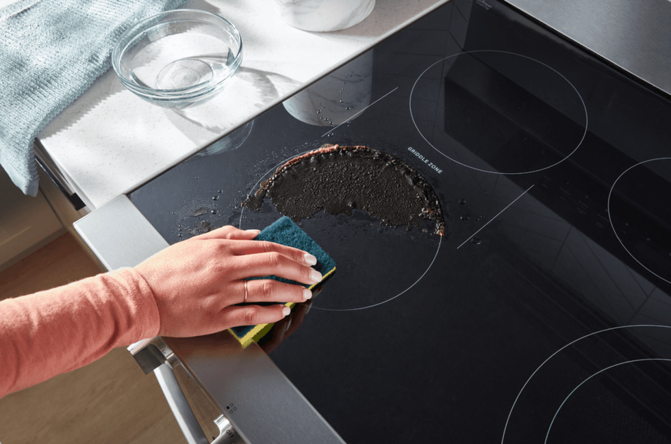 A hand wipes mess off the top of a electric oven range with a yellow and green sponge.