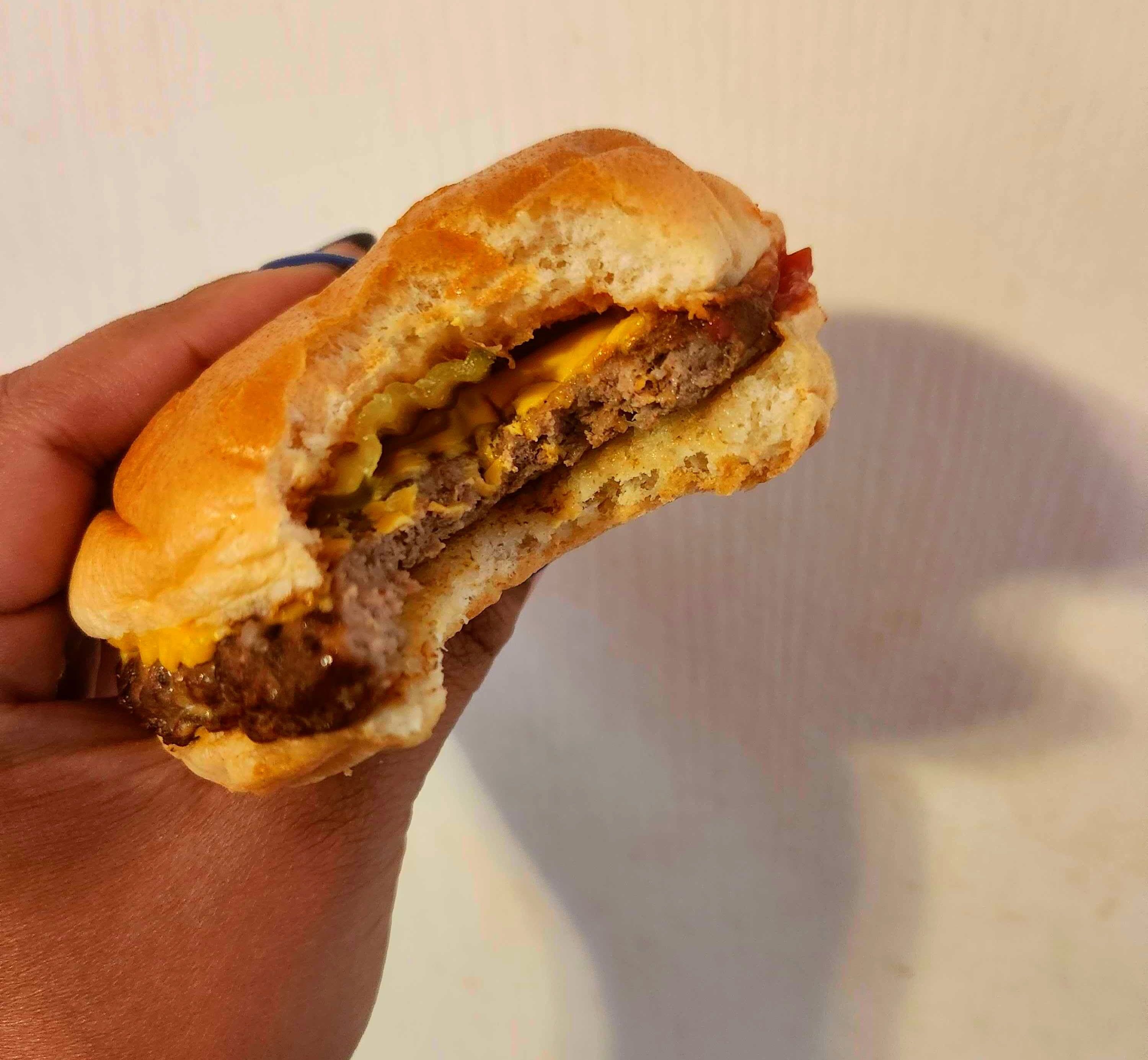 A hand with blue nail polish holding a hamburger against a white wall.