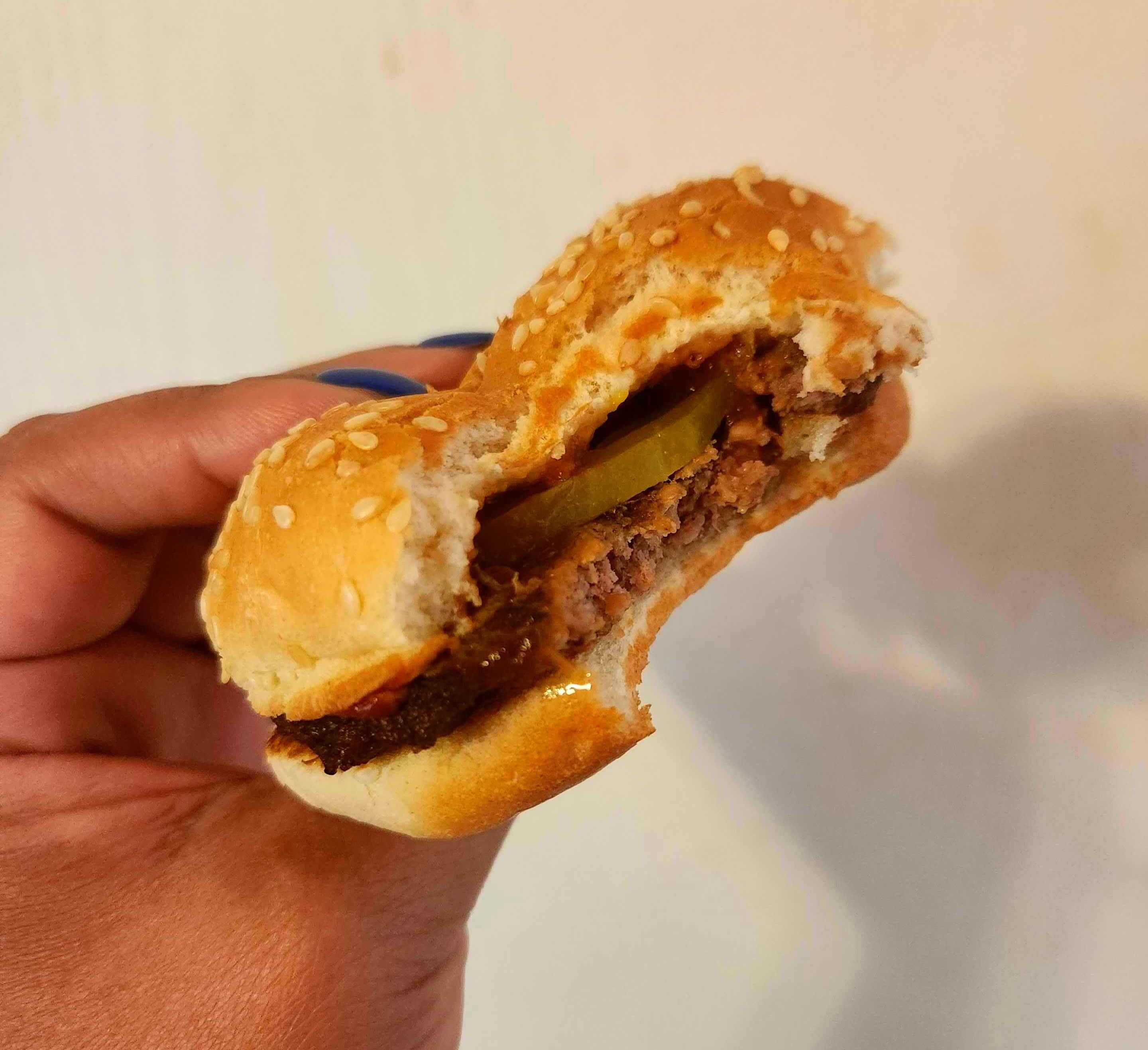 A hand with blue nail polish holding a hamburger against a white wall.
