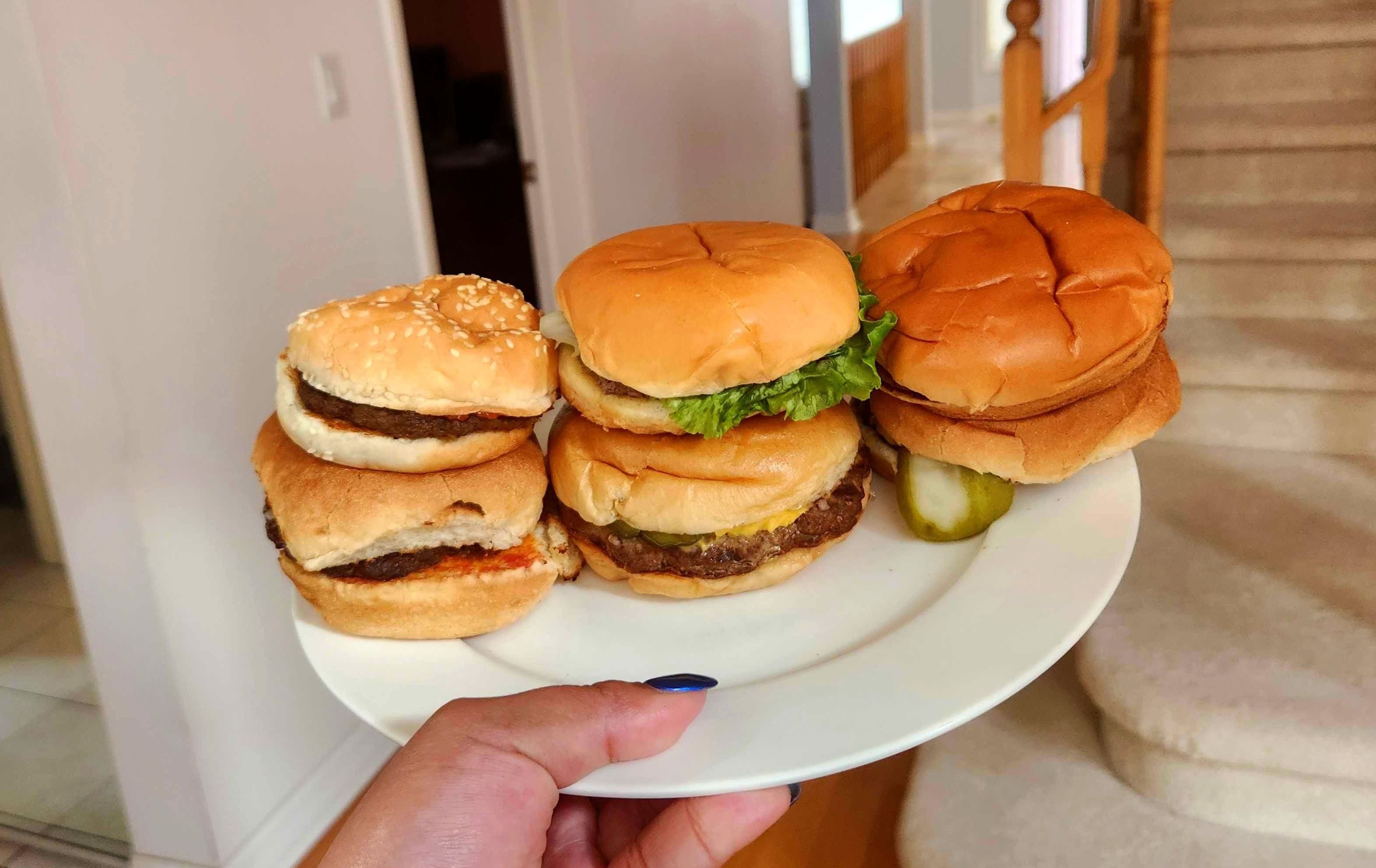 A hand with blue nail polish holding a white plate with six fast food burgers on it.