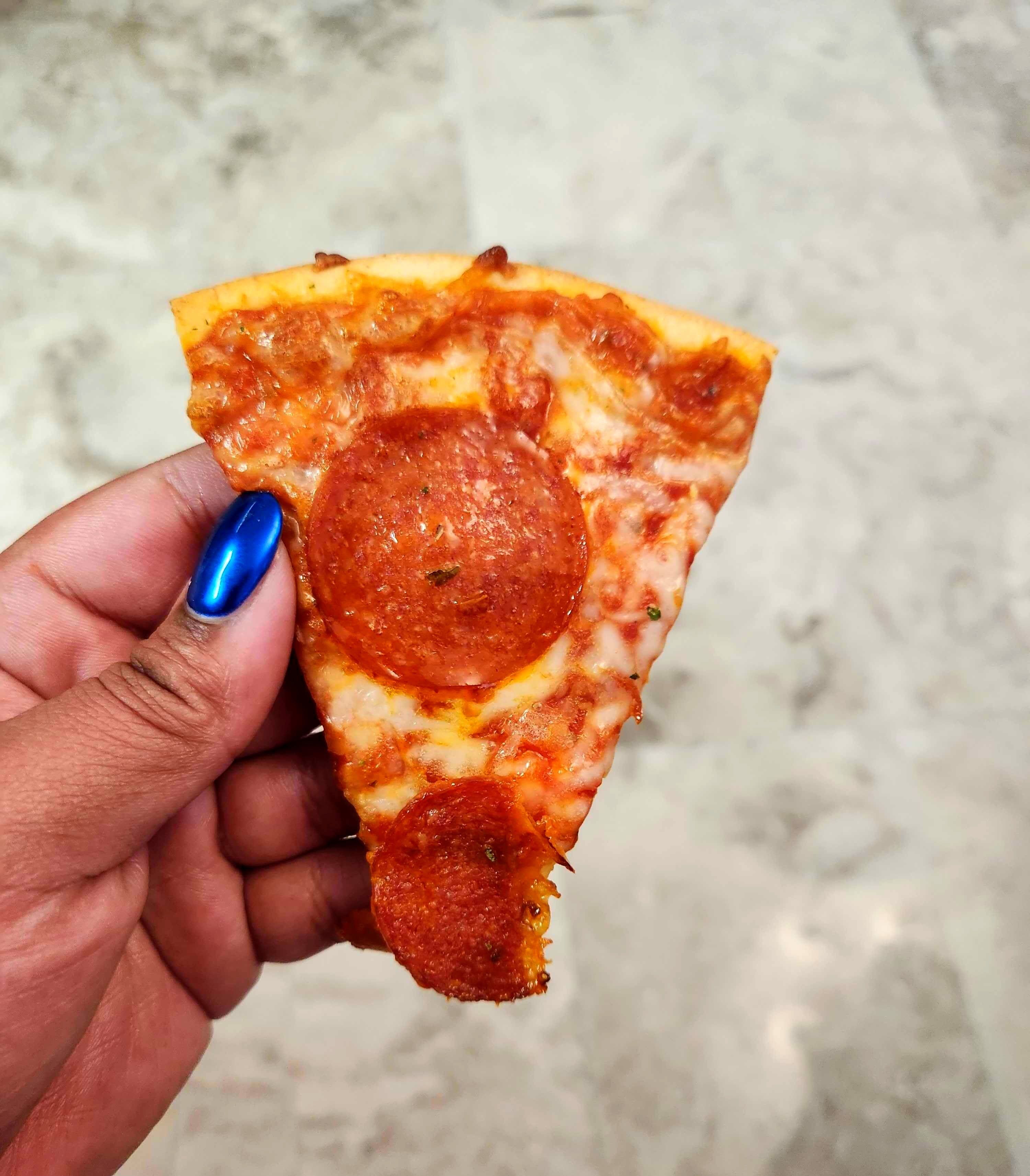 A hand with blue nails holding a slice of pizza against a tile background.