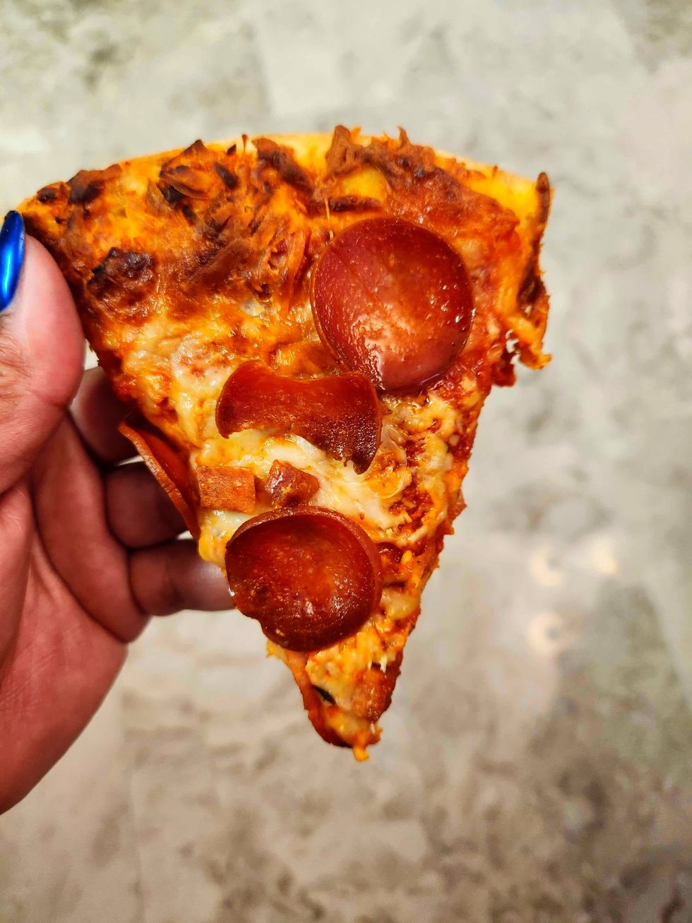 A hand with blue nails holding a slice of pizza against a tile background.