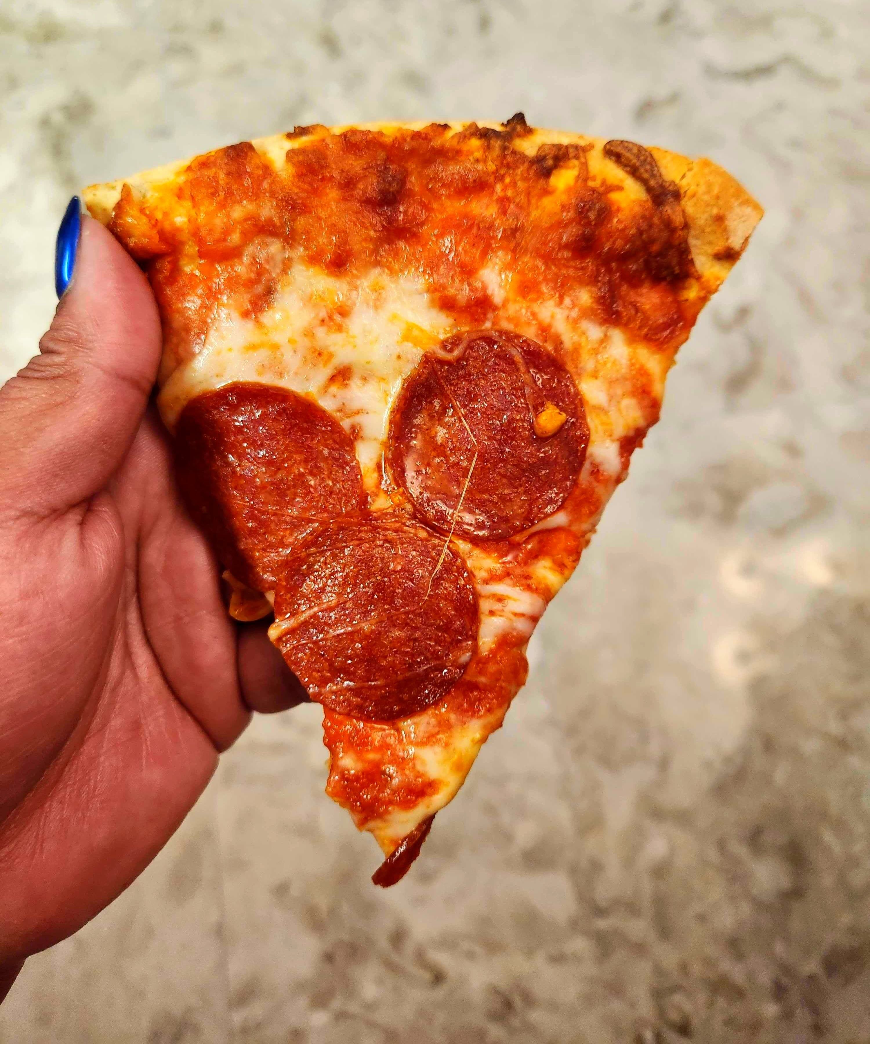 A hand with blue nails holding a slice of pizza against a tile background.