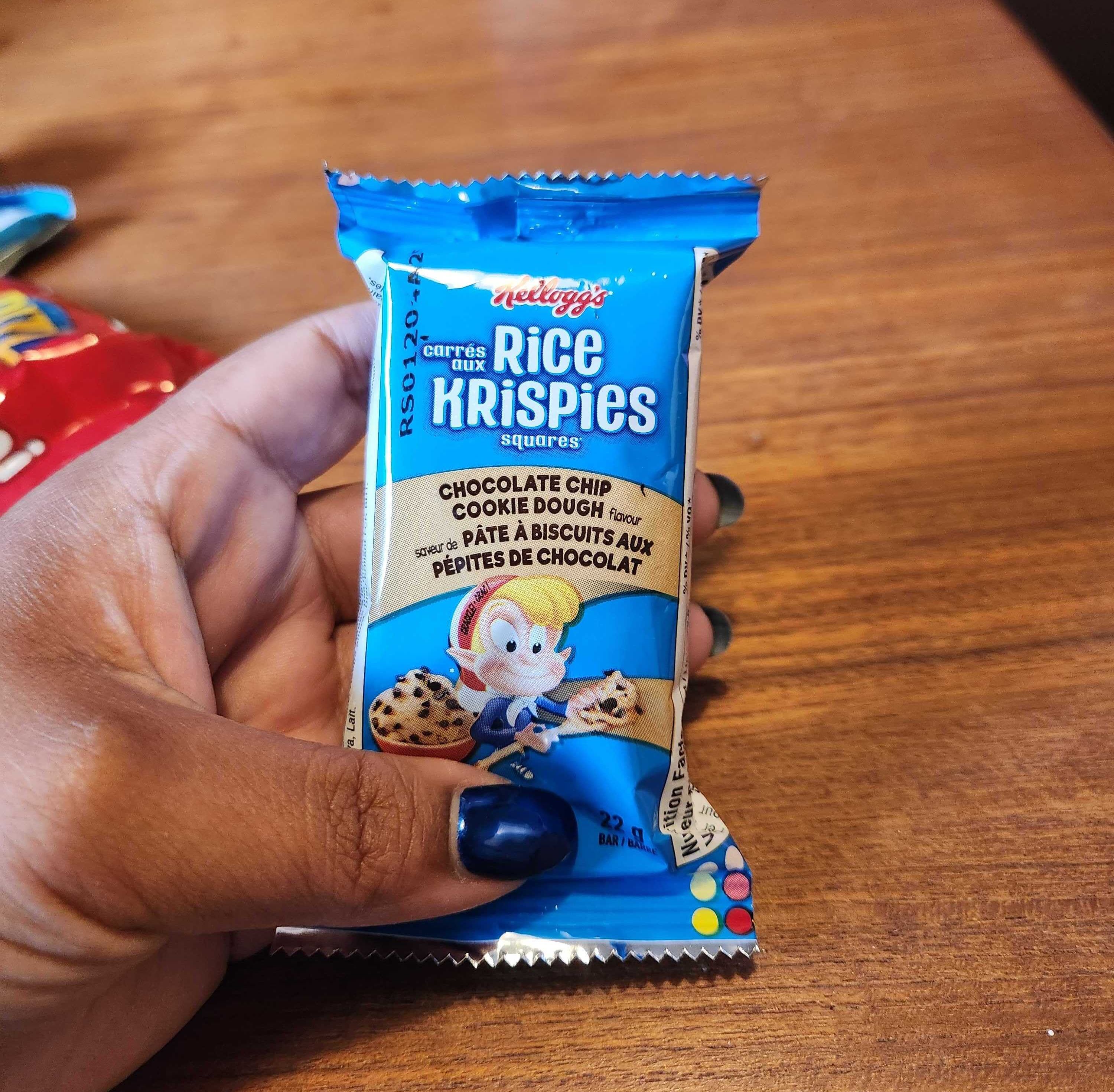 A hand with blue nails holding a snack with blue wrapping against a brown wooden table.