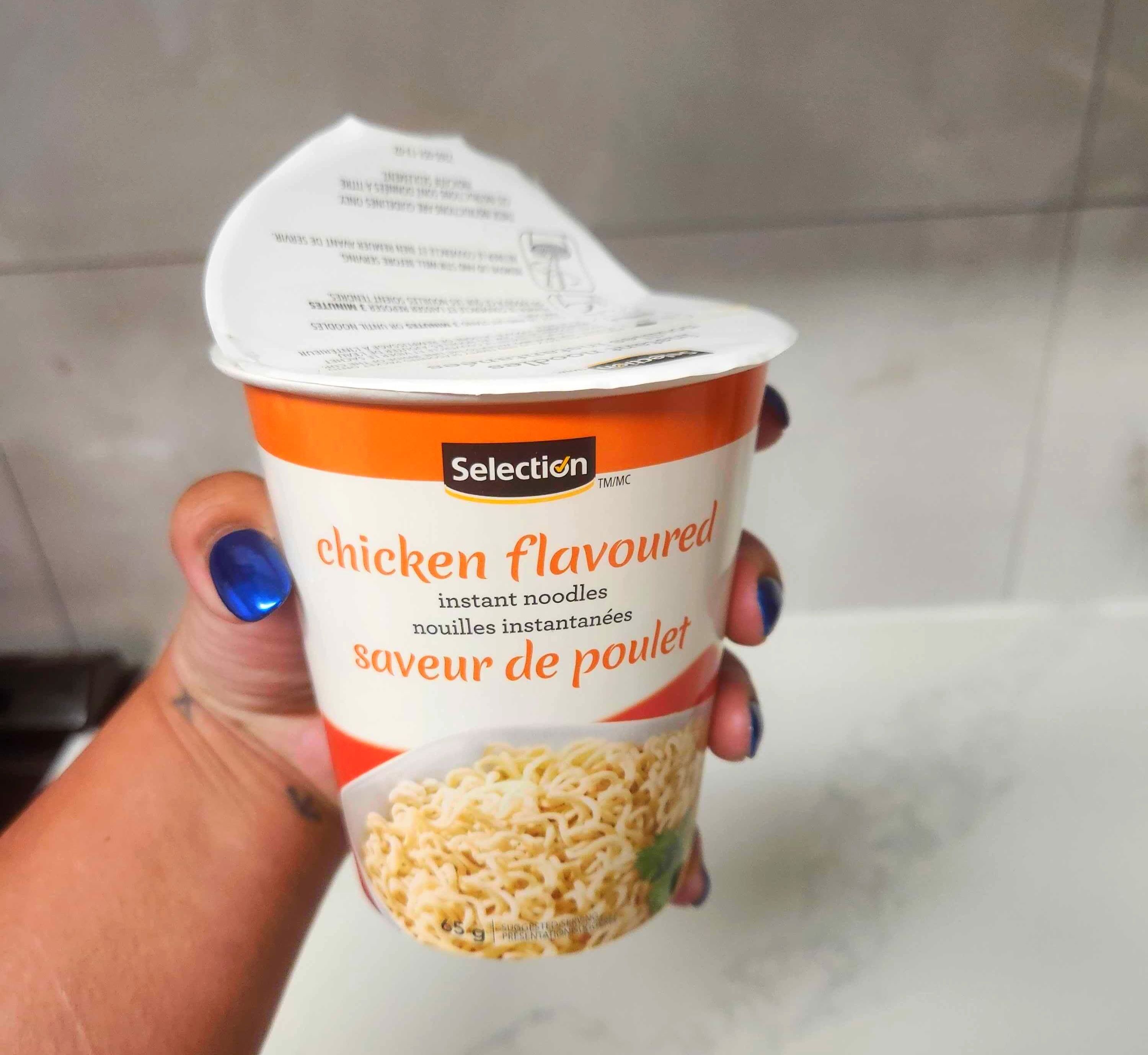 A hand with blue nails holding a white container of soup against a marble counter.
