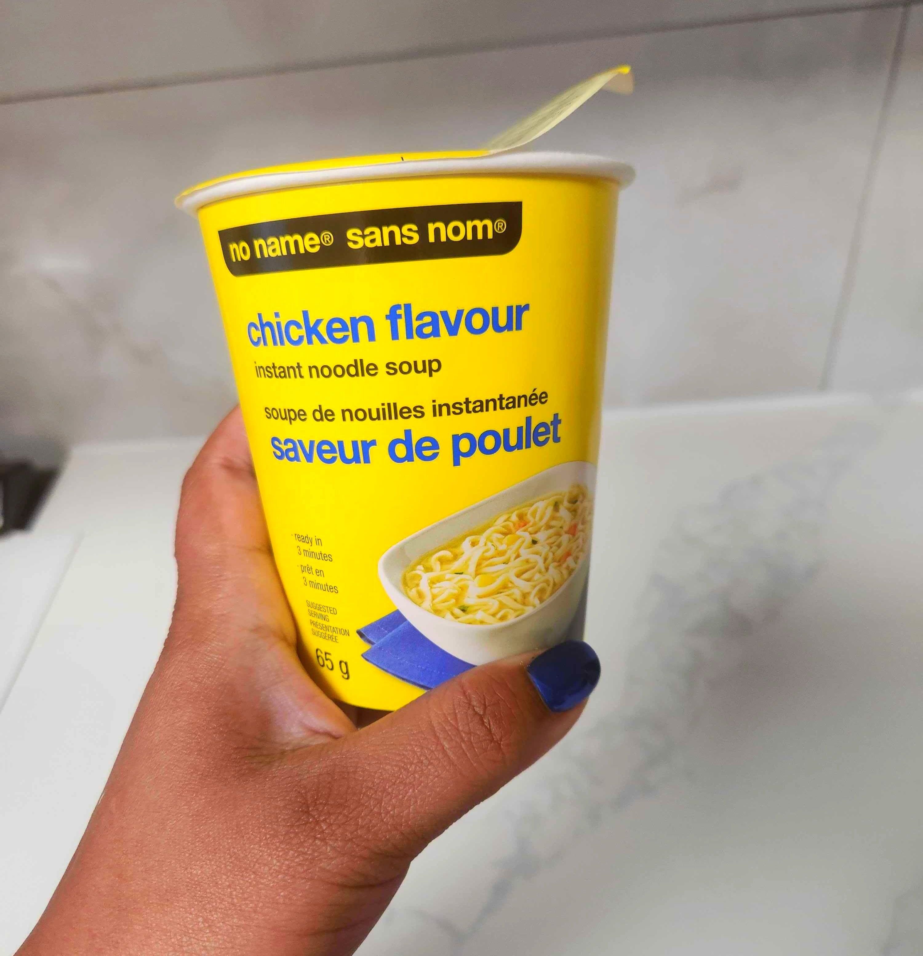 A hand with blue nails holding a yellow container of soup against a marble counter.