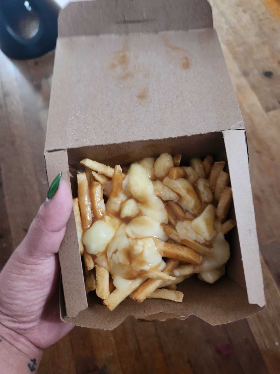 A hand with green nail polish and an anchor tattoo on the wrist holding a brown box of poutine.