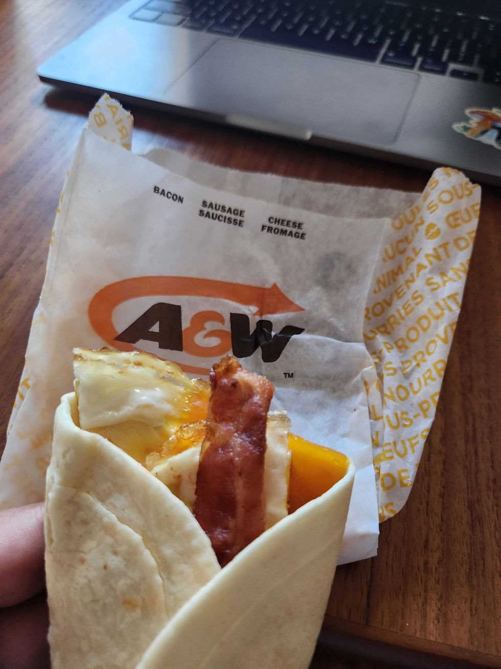 A hand with green nails holding a breakfast wrap in front of a brown wooden table and a white A&W paper bag.