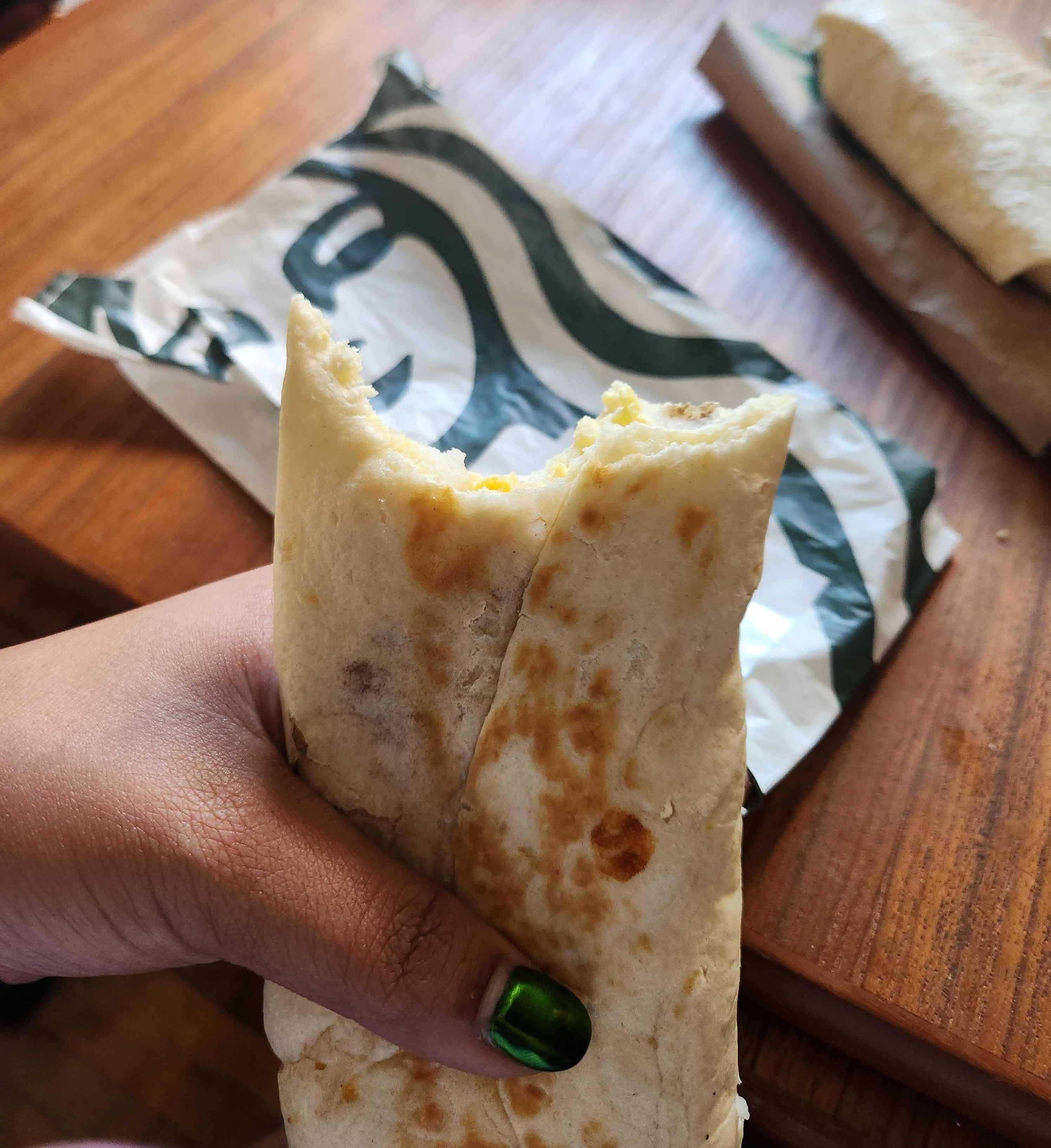 A hand with green nails holding a breakfast wrap with a bite taken out of it in front of a brown wooden table and a white and green Starbucks paper bag.