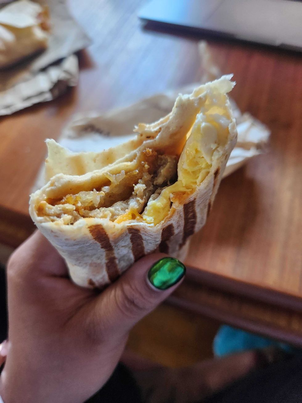 A hand with green nails holding a breakfast wrap with a bite taken out of it in front of a brown wooden table.