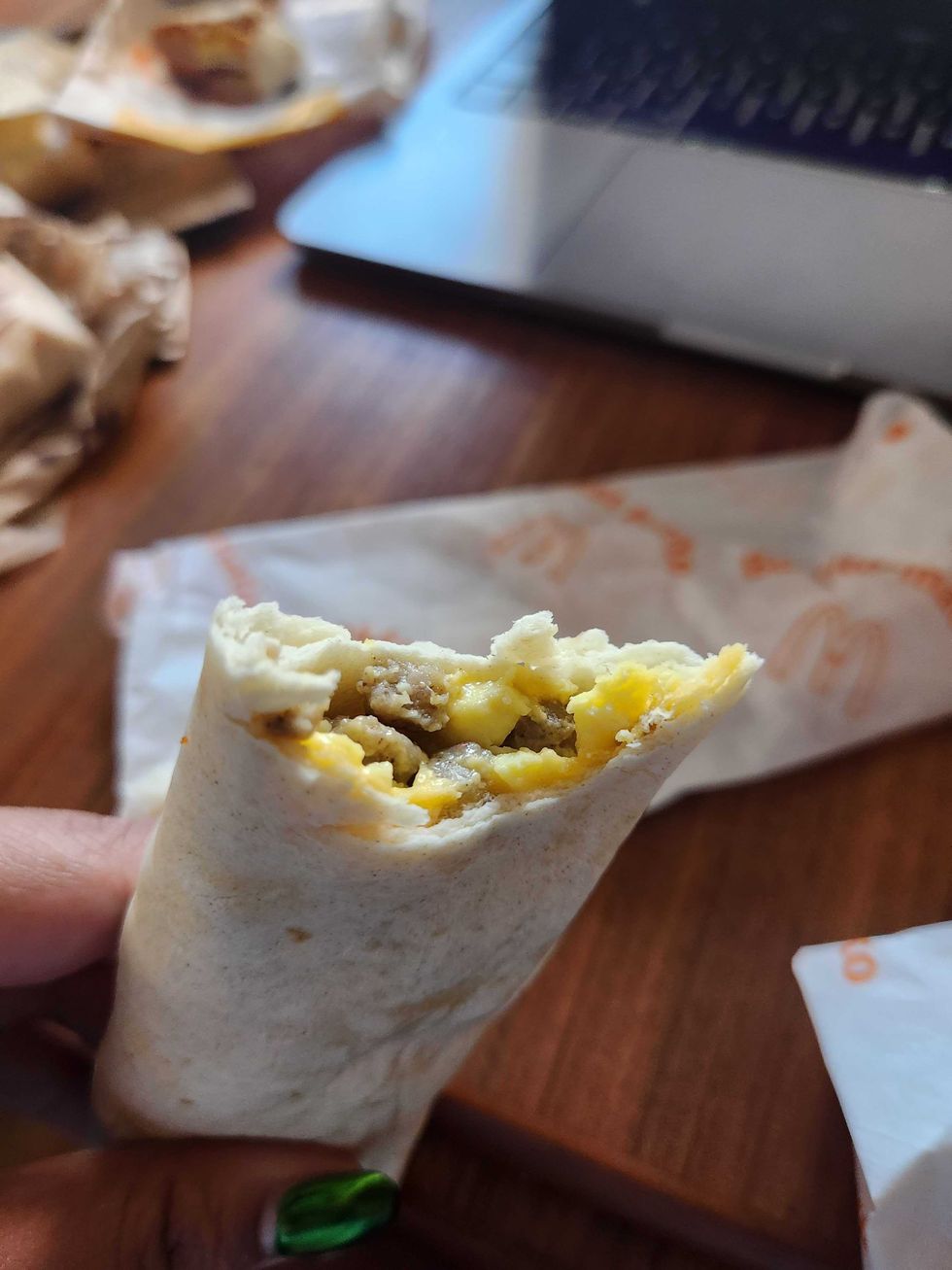 A hand with green nails holding a breakfast wrap with a bite taken out of it in front of a brown wooden table.