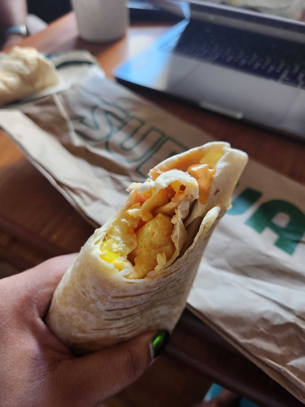 A hand with green nails holding a breakfast wrap with a bite taken out of it in front of a brown wooden table.