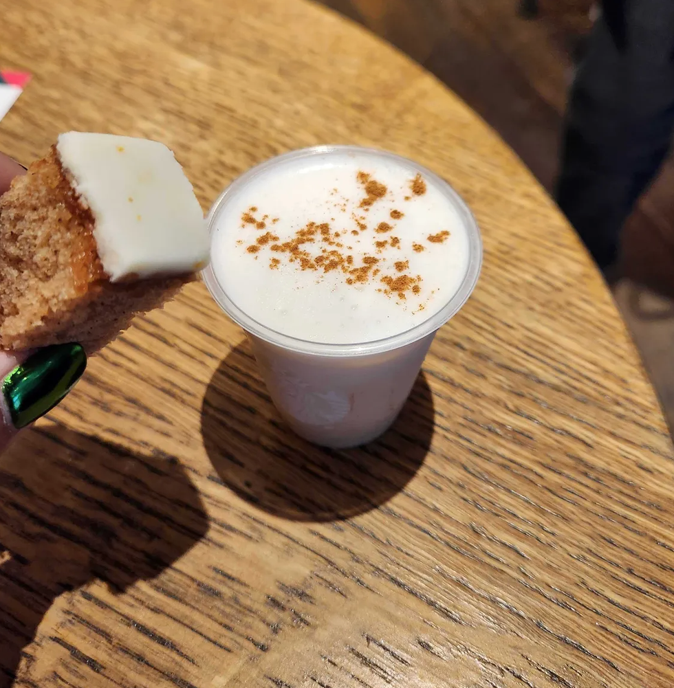 A hand with green nails holding a brown slice of cake with white frosting on a table with an iced white drink with white foam and brown dust on top.