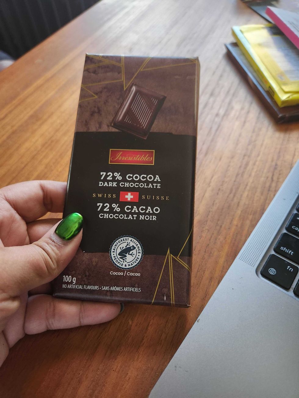 A hand with green nails holding a chocolate bar with a dark brown and black wrapper on a brown wooden table.