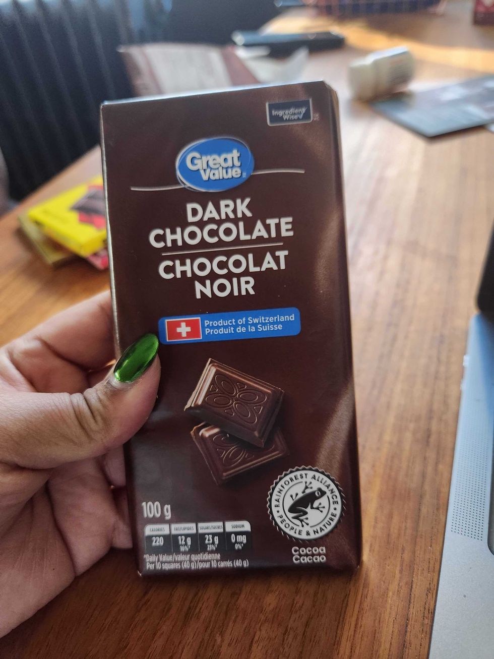 A hand with green nails holding a chocolate bar with a dark brown wrapper on a brown wooden table.