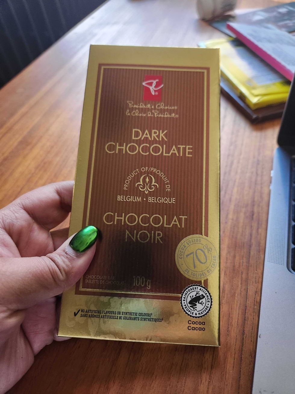 A hand with green nails holding a chocolate bar with a gold and brown wrapper on a brown wooden table.