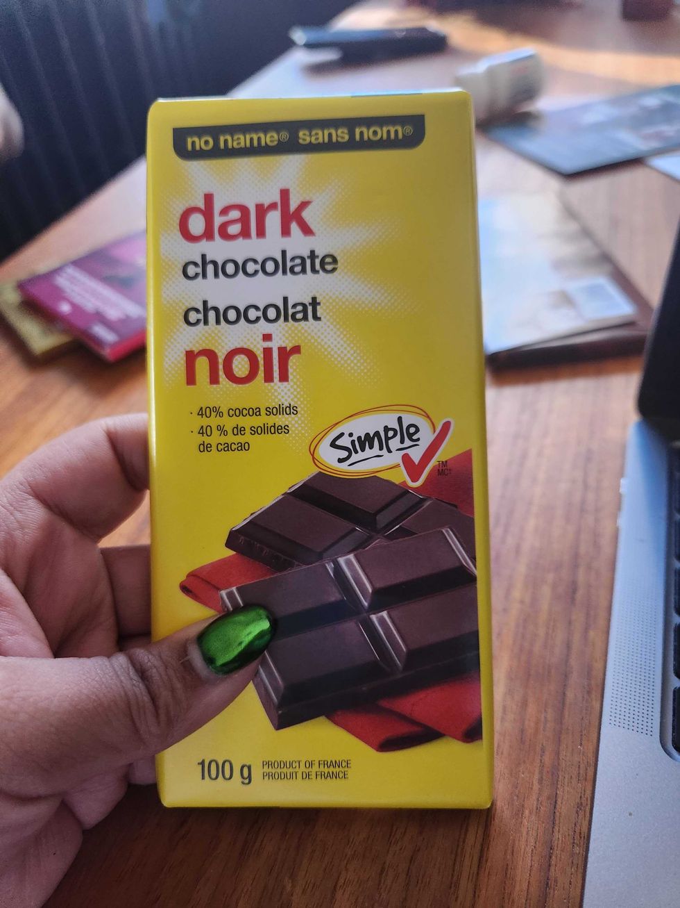 A hand with green nails holding a chocolate bar with a yello wrapper on a brown wooden table.