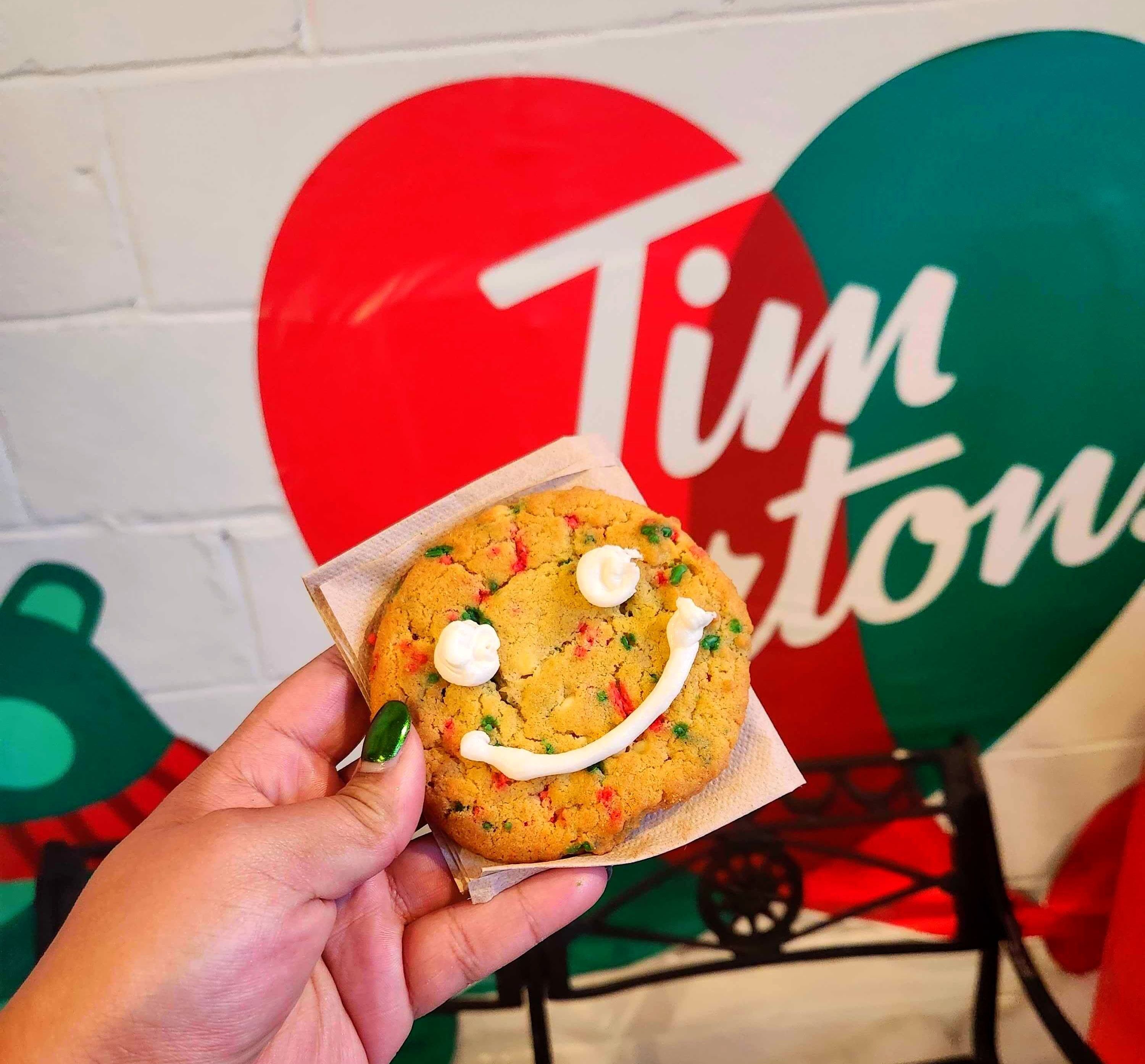 A hand with green nails holding a cookie with green and red sprinkles and a white iced smiley face.