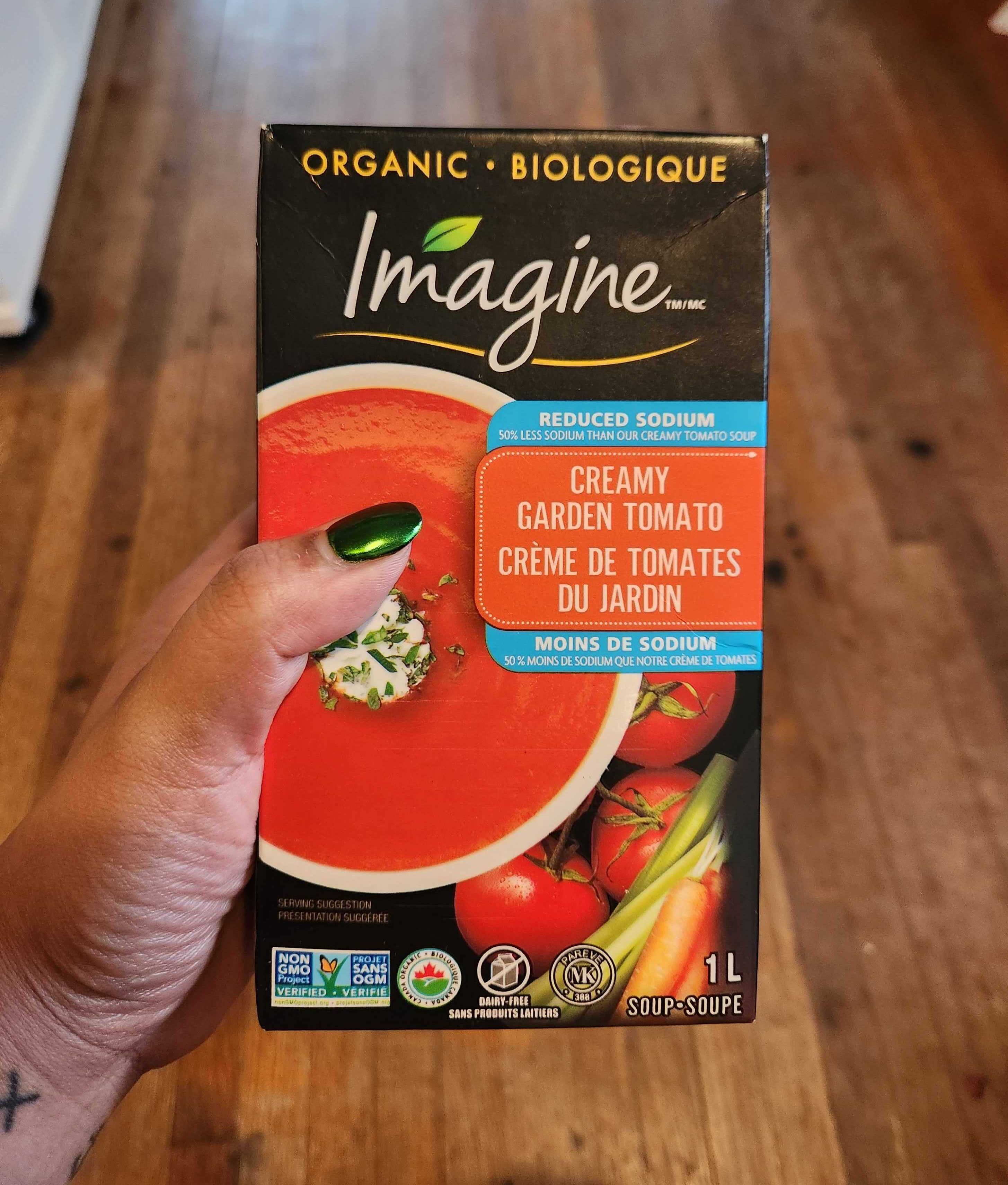 A hand with tattoos and green nail polish holding a container of soup.