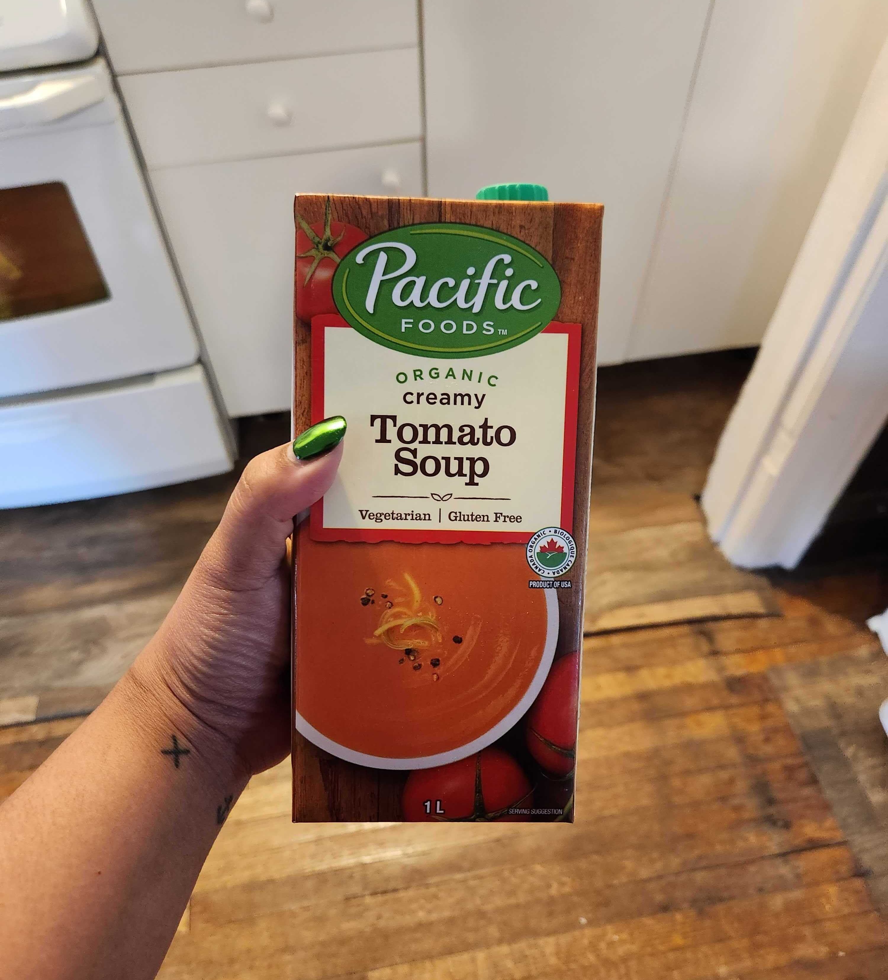 A hand with tattoos and green nail polish holding a container of soup.
