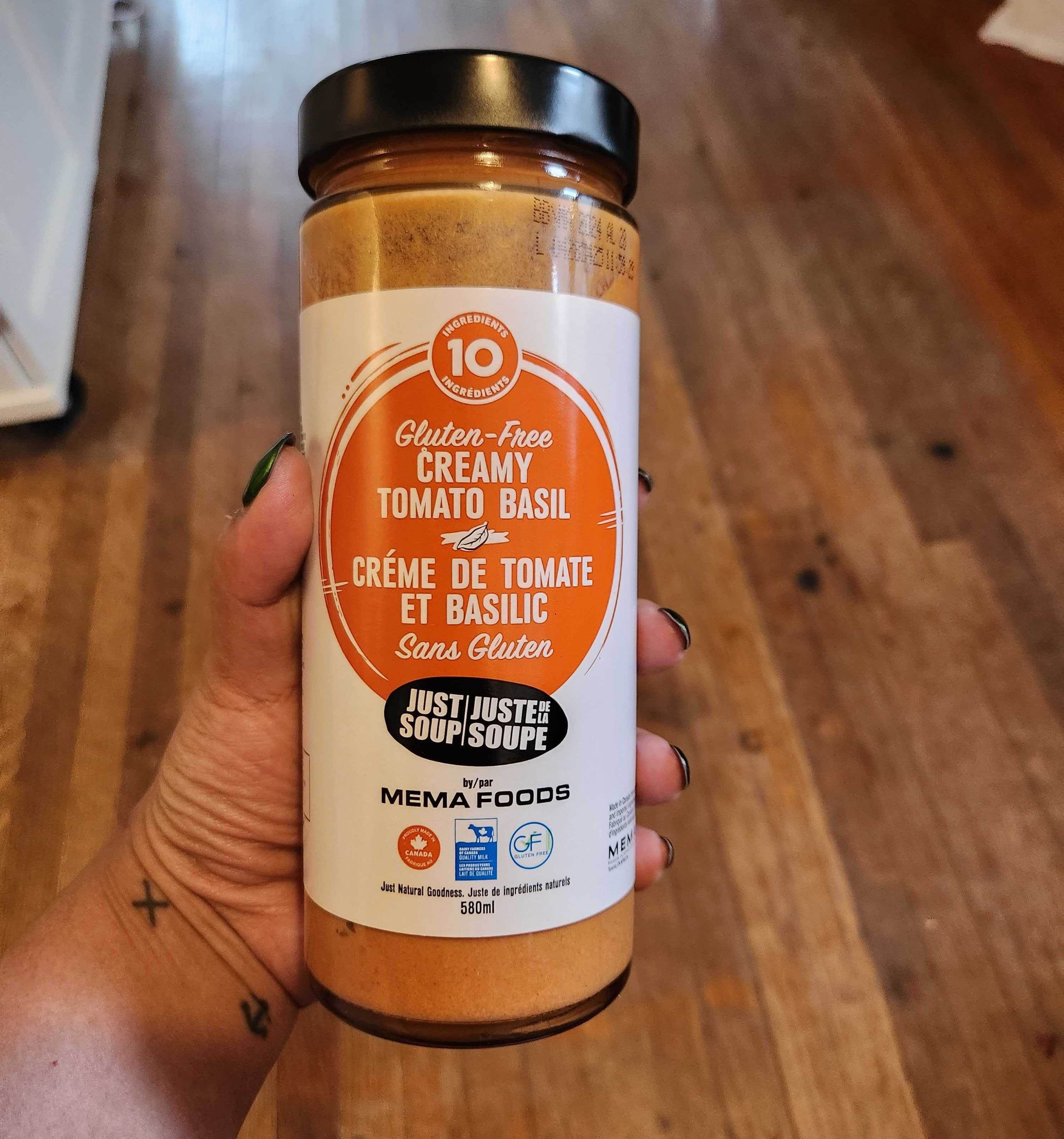 A hand with tattoos and green nail polish holding a jar of soup.