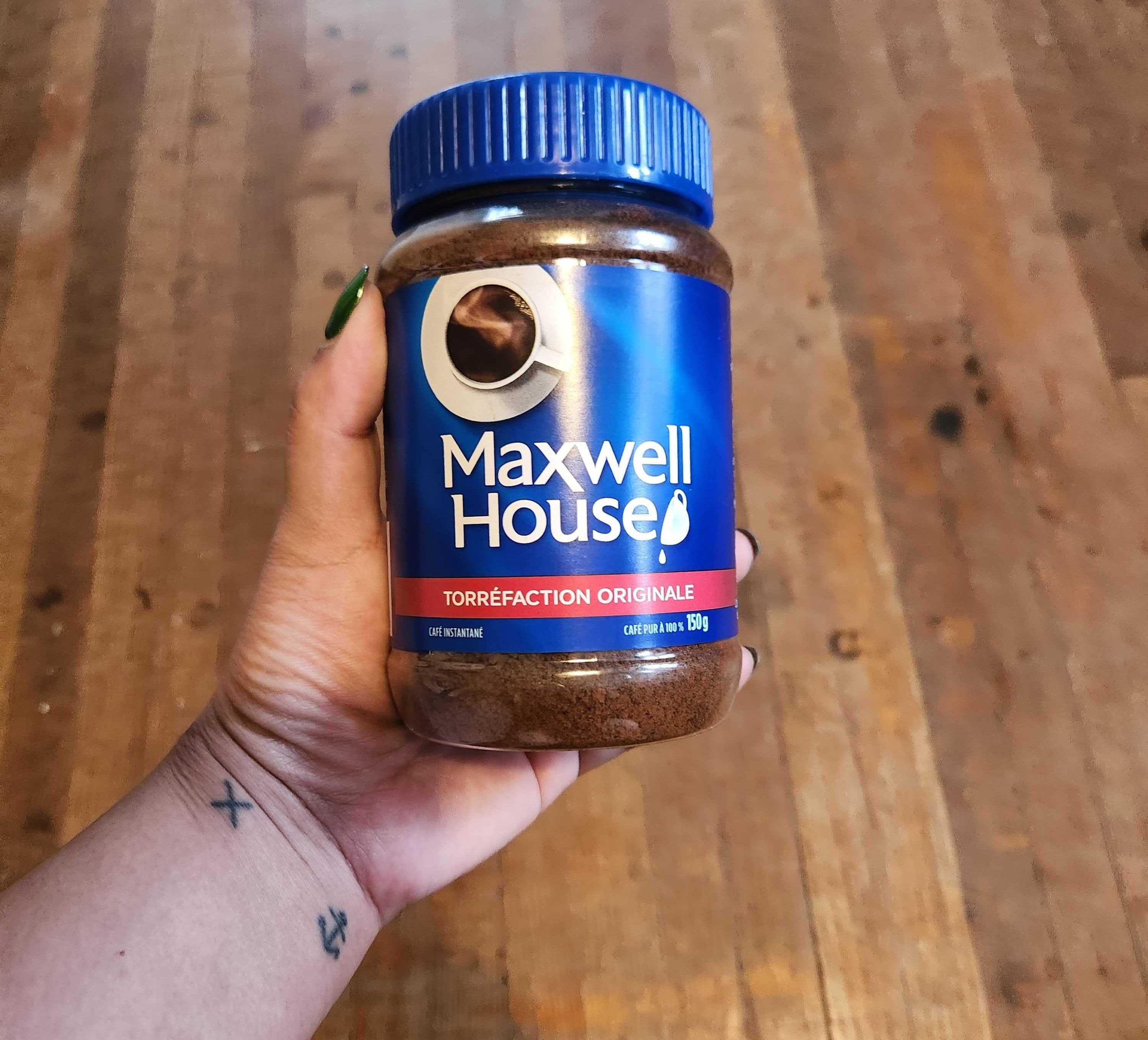 A hand with tattoos and green nails holding a coffee jar with a blue label and blue lid.