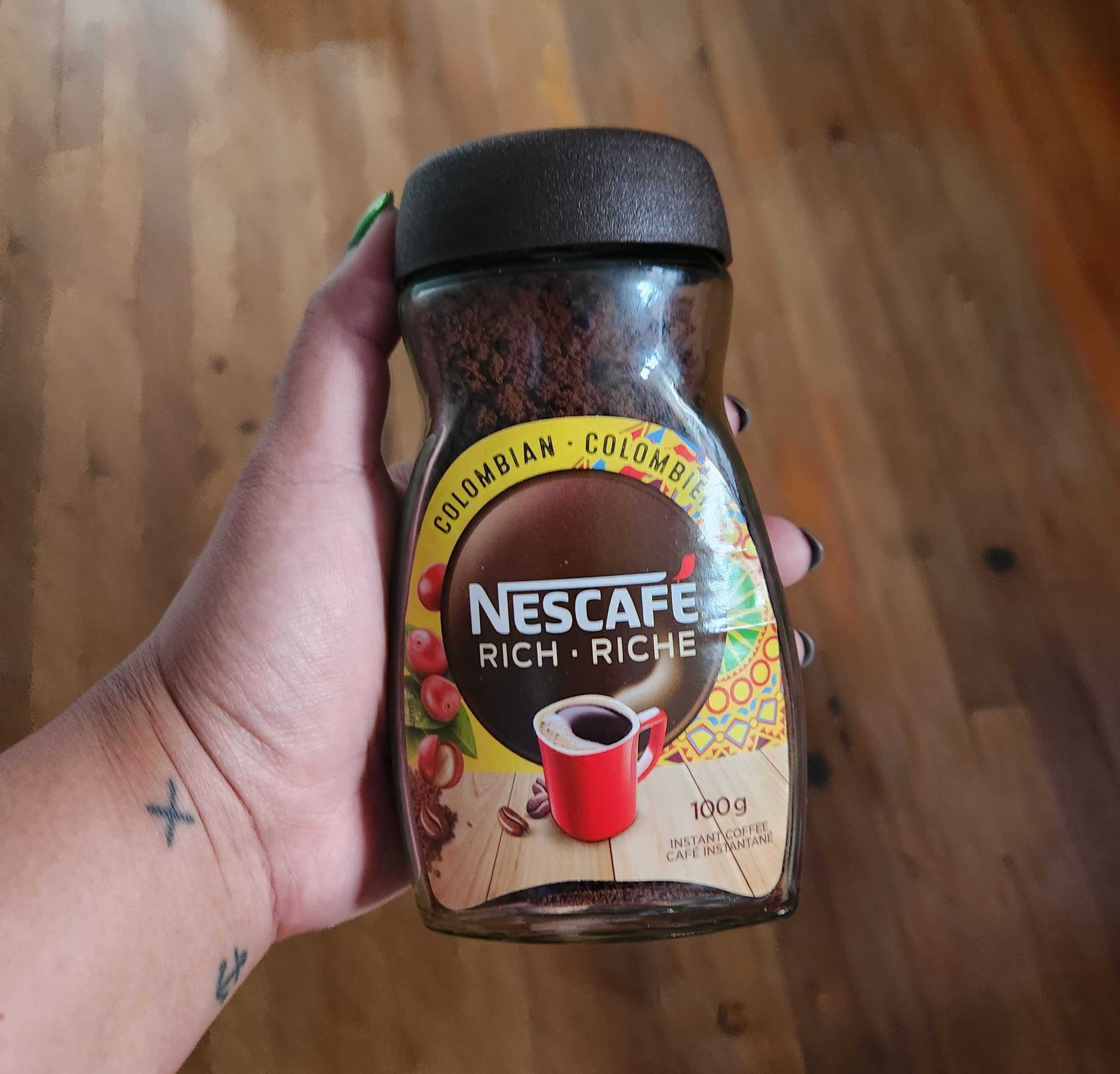 A hand with tattoos and green nails holding a coffee jar with a brown label and black lid.