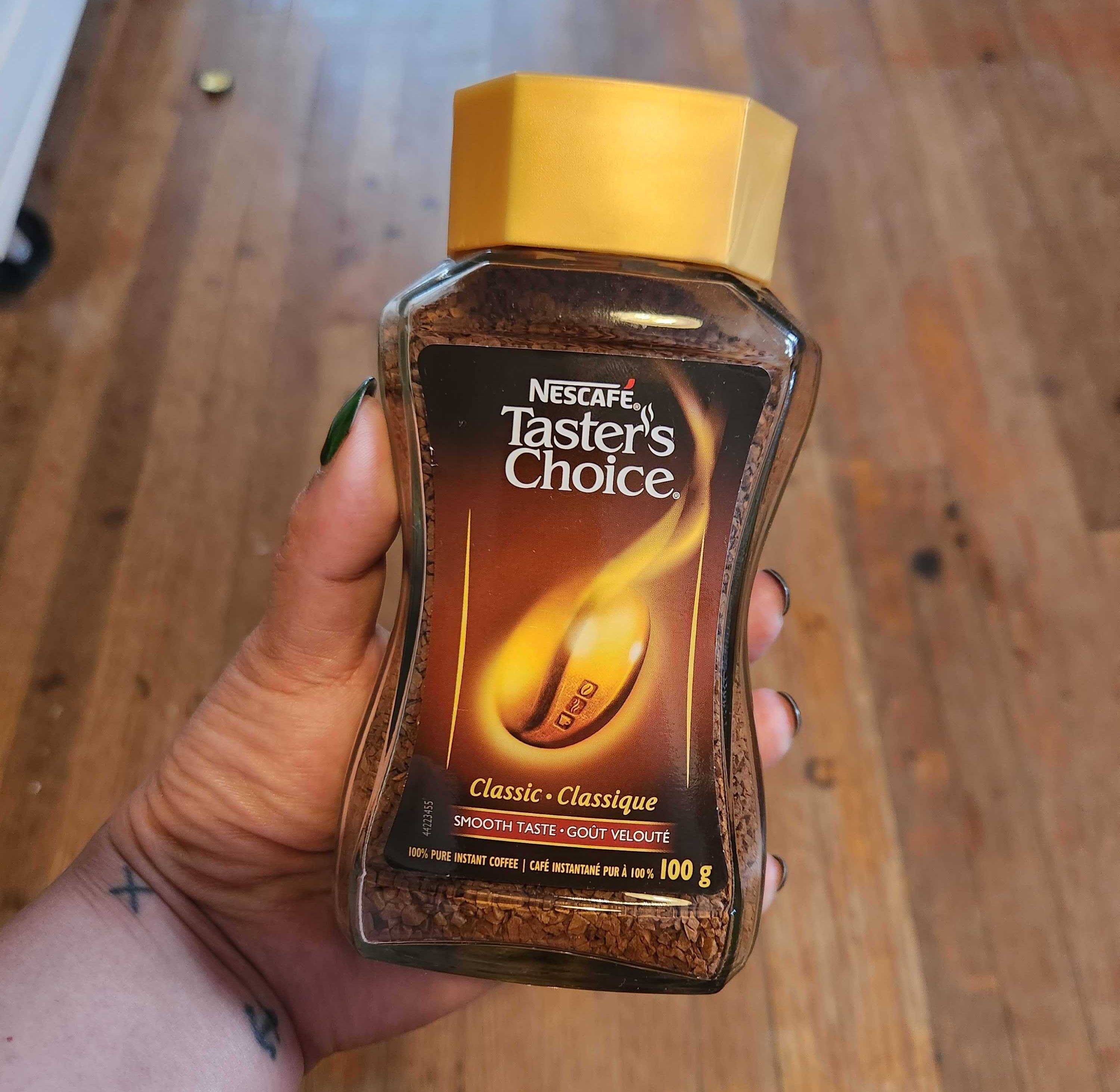 A hand with tattoos and green nails holding a coffee jar with a brown label and yellow lid.