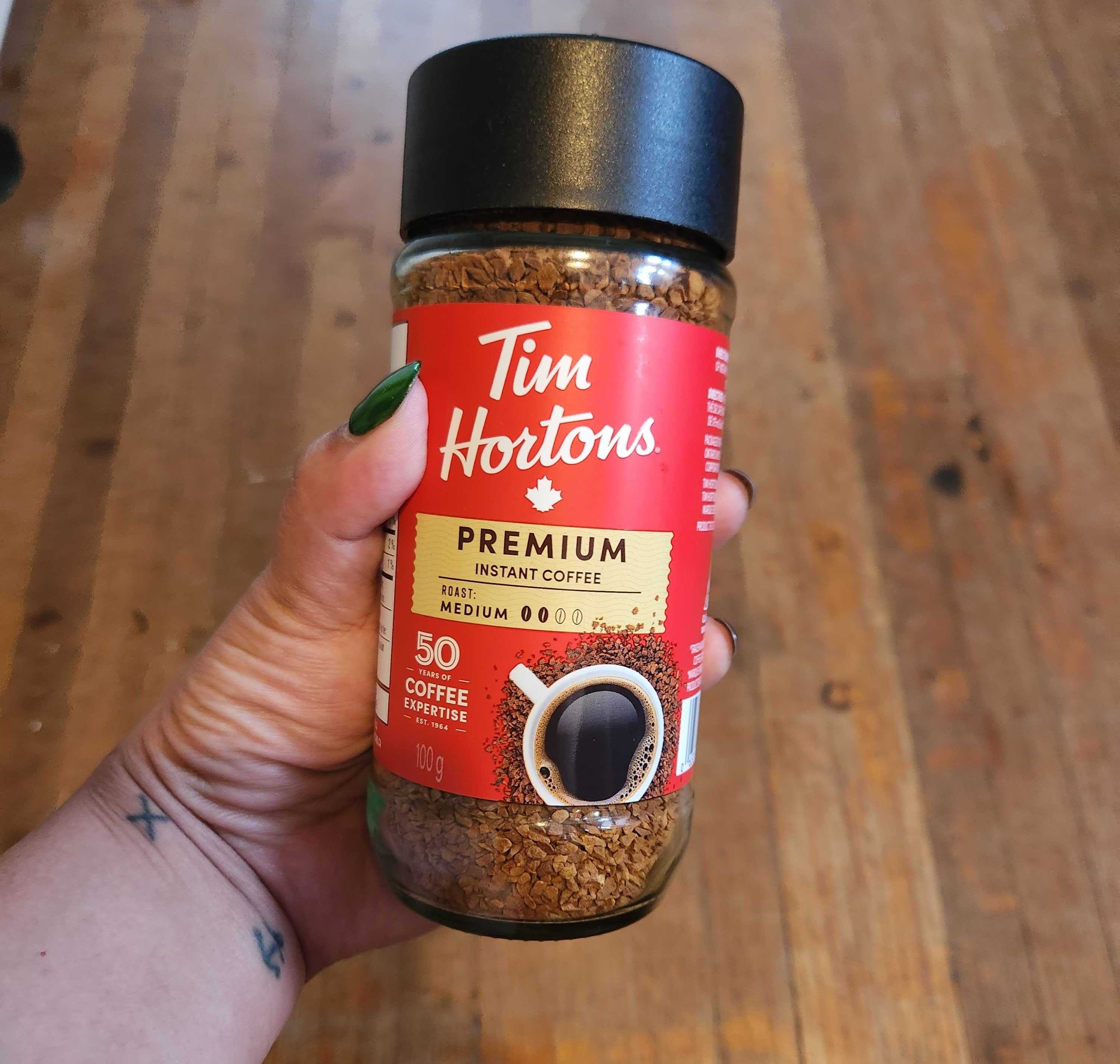 A hand with tattoos and green nails holding a coffee jar with a red label and black lid.