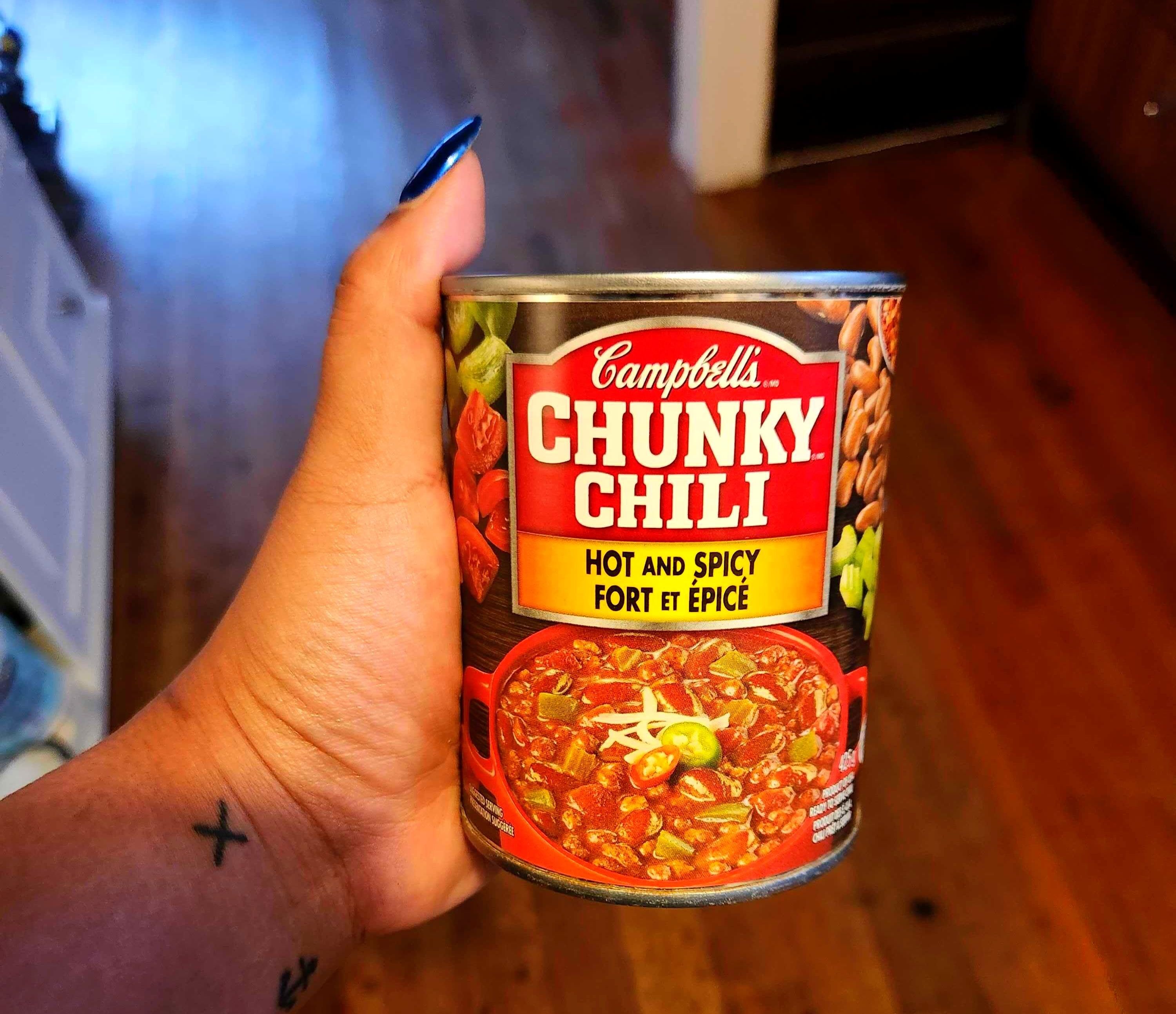 A hand with tattoos holding a tin with a red and brown label against a brown floor.