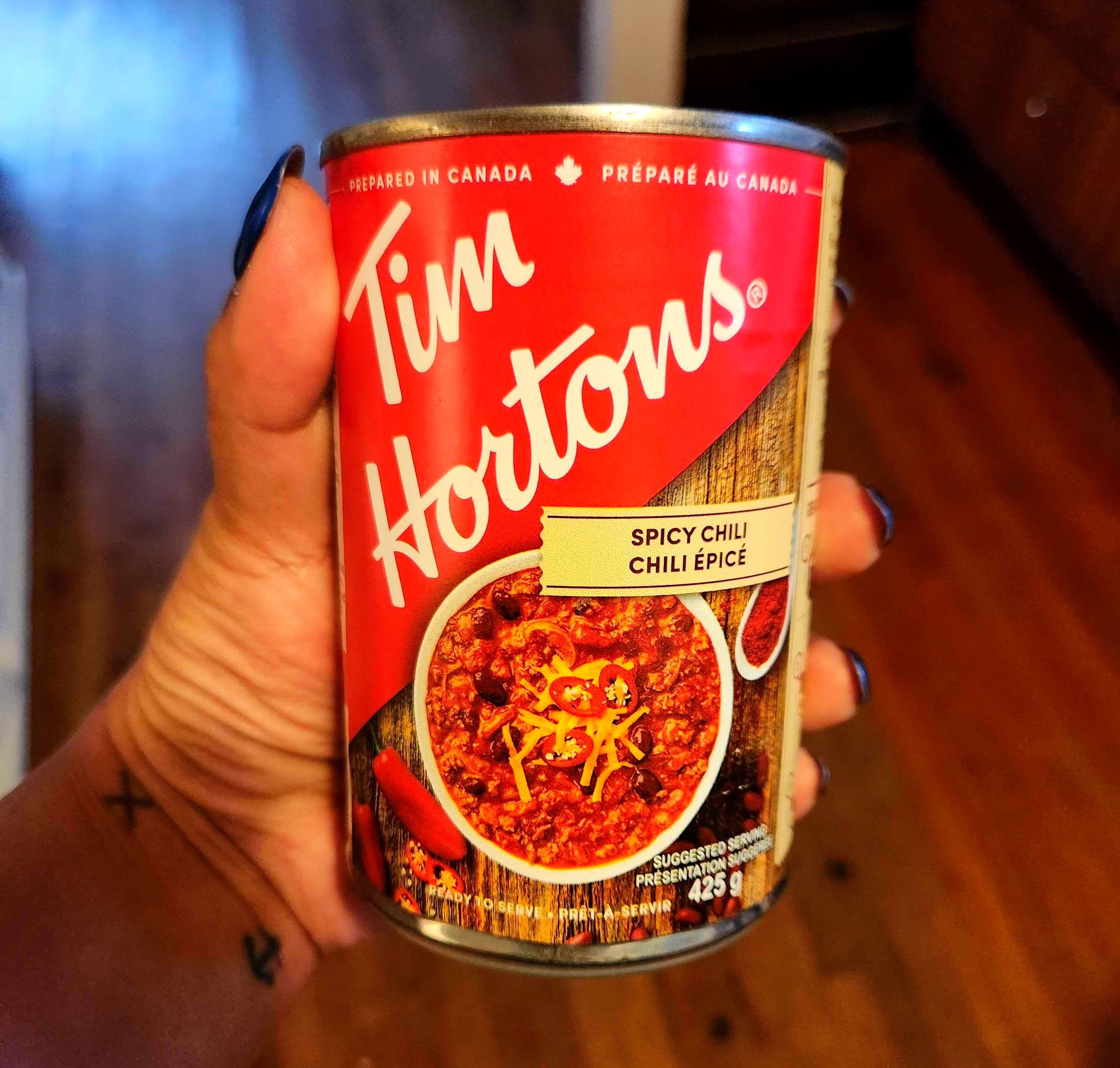A hand with tattoos holding a tin with a red label against a brown floor.