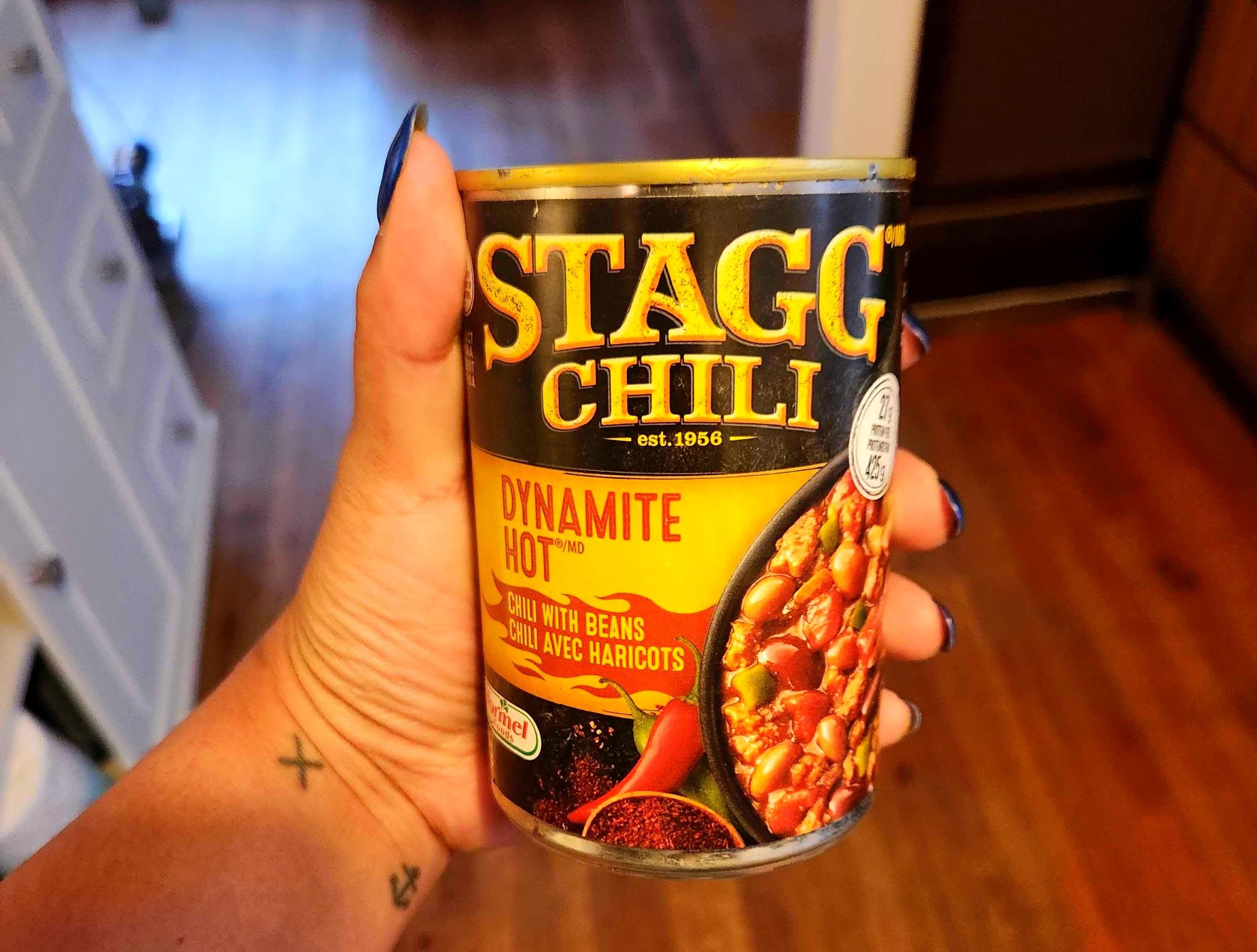 A hand with tattoos holding a tin with a yellow ad black label against a brown floor.