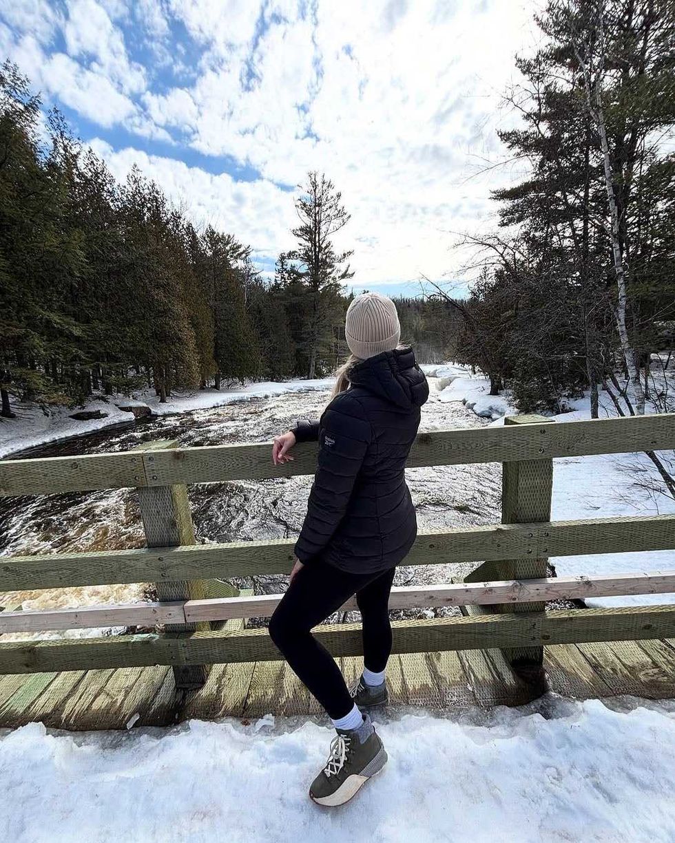 A hiker on a bridge looks out over a river flowing through the snow.