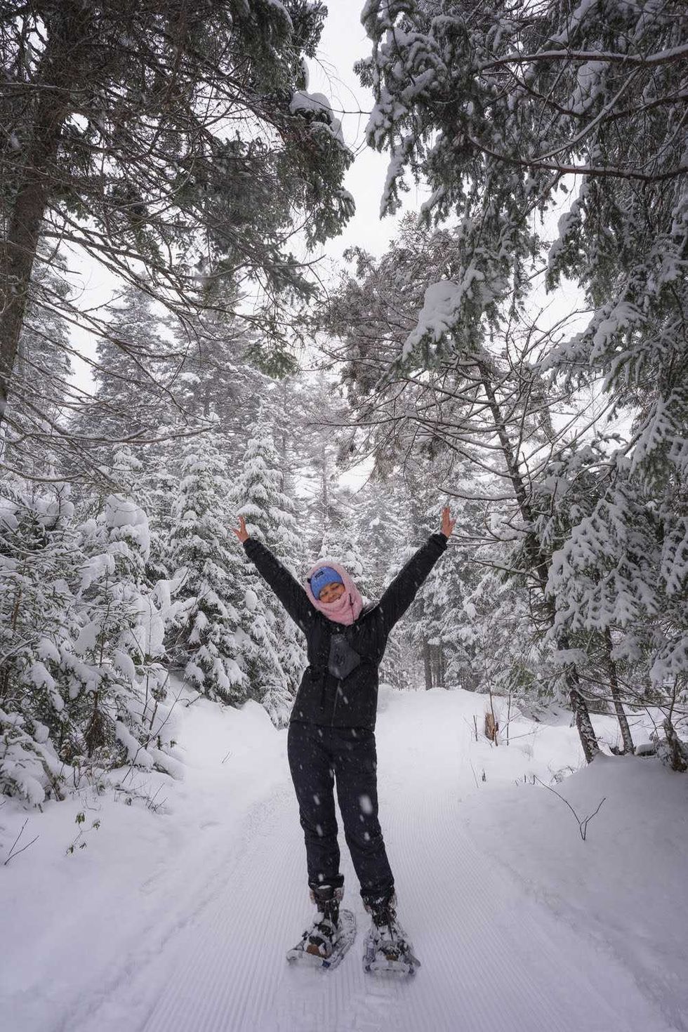 A hiker wearing snowshoes with thier arms raised on a forest trail.