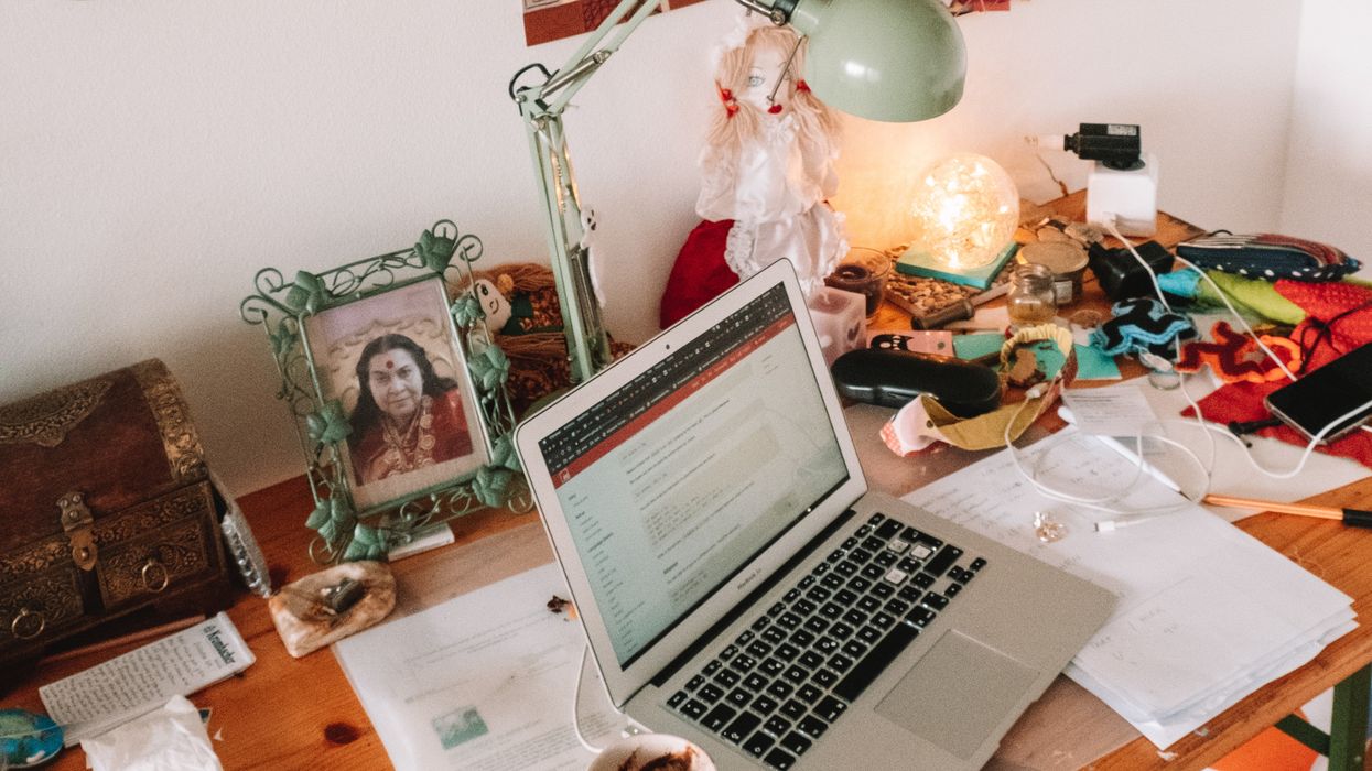 A laptop on a desk, surrounded by papers, a picture frame, a lamp and other belongings.