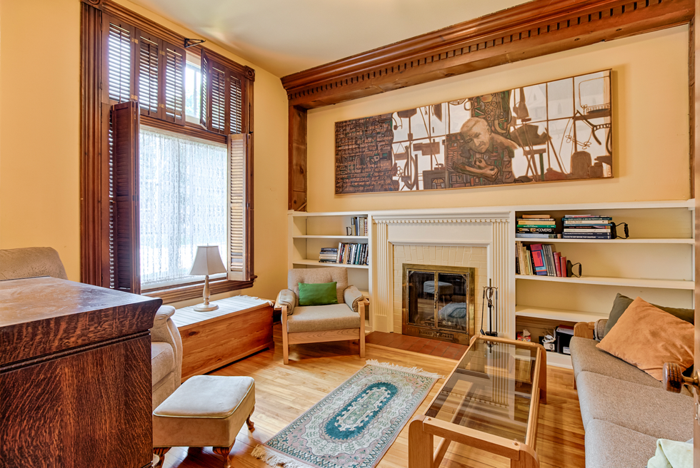 A living room with a fireplace, sofa, chairs, table, booked and a large painting hanging on the wall.