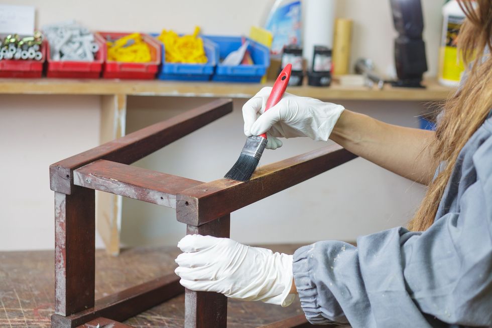 A long-haired person wearing latex gloves and a paint smock stains the leg of a stool using a paintbrush.