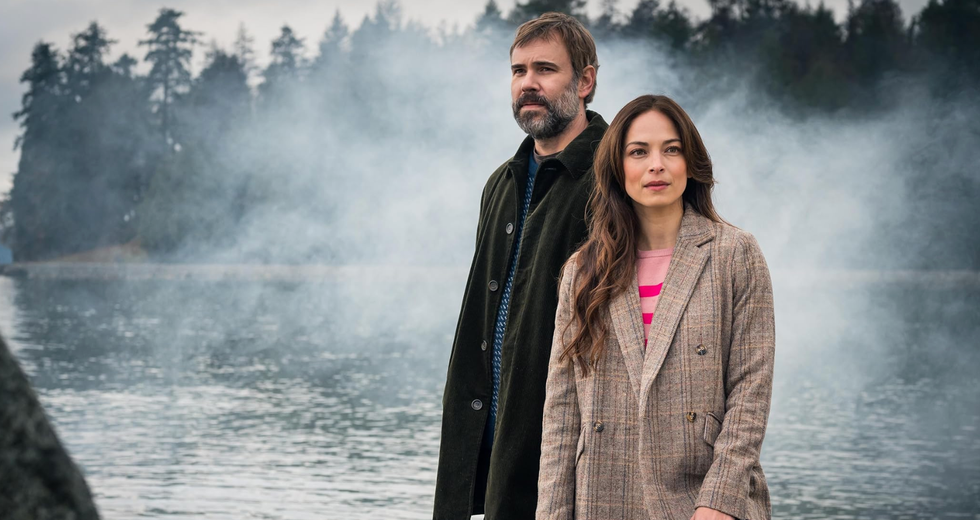 A man and woman in front of a body of water, trees and fog.