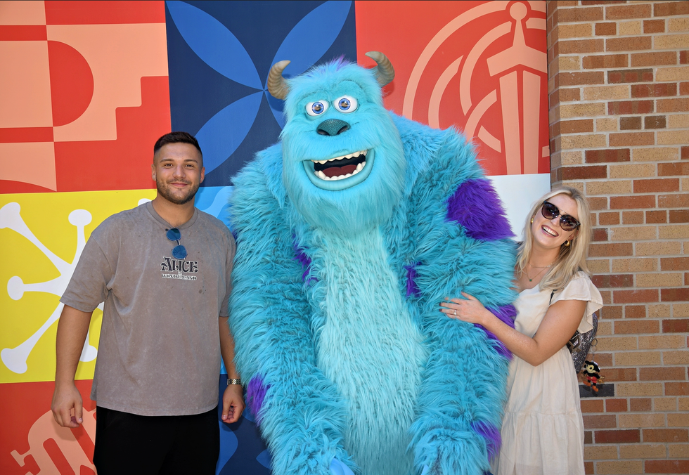 A man and woman smiling while posing with Sulley at Disney World in front of a colourful Pixar-themed backdrop.