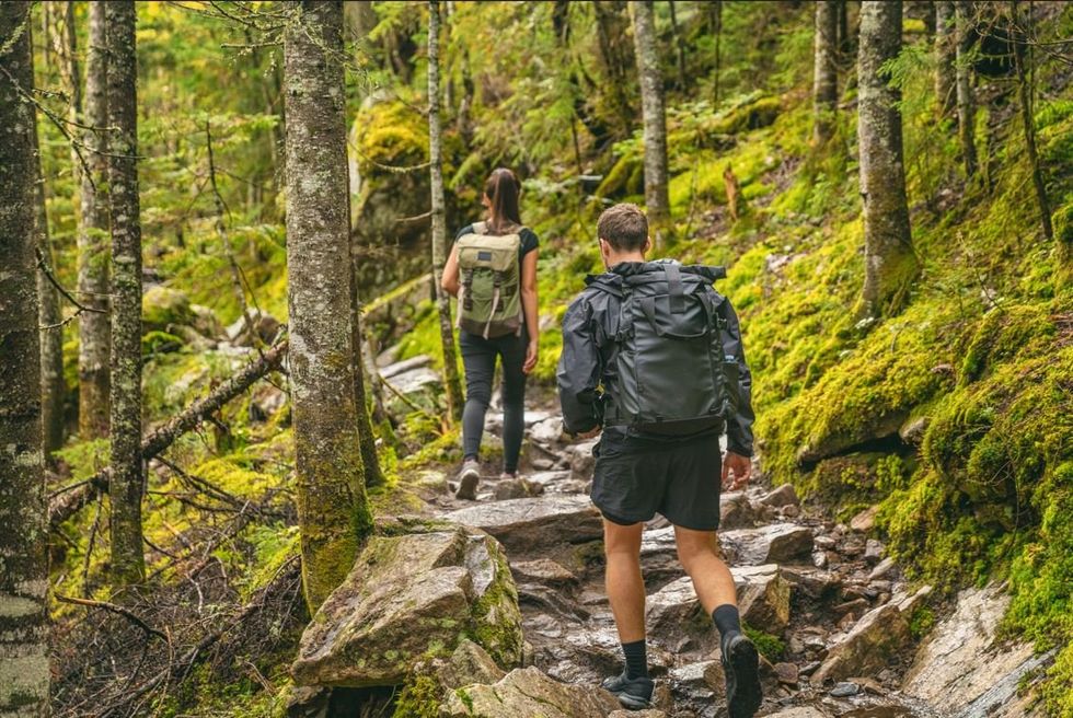 A man and woman walk along a rocky hiking trail in a forest.