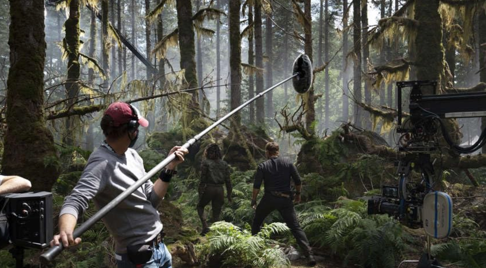 A man holding a microphone above two people walking into a forest with a camera filming on the side.