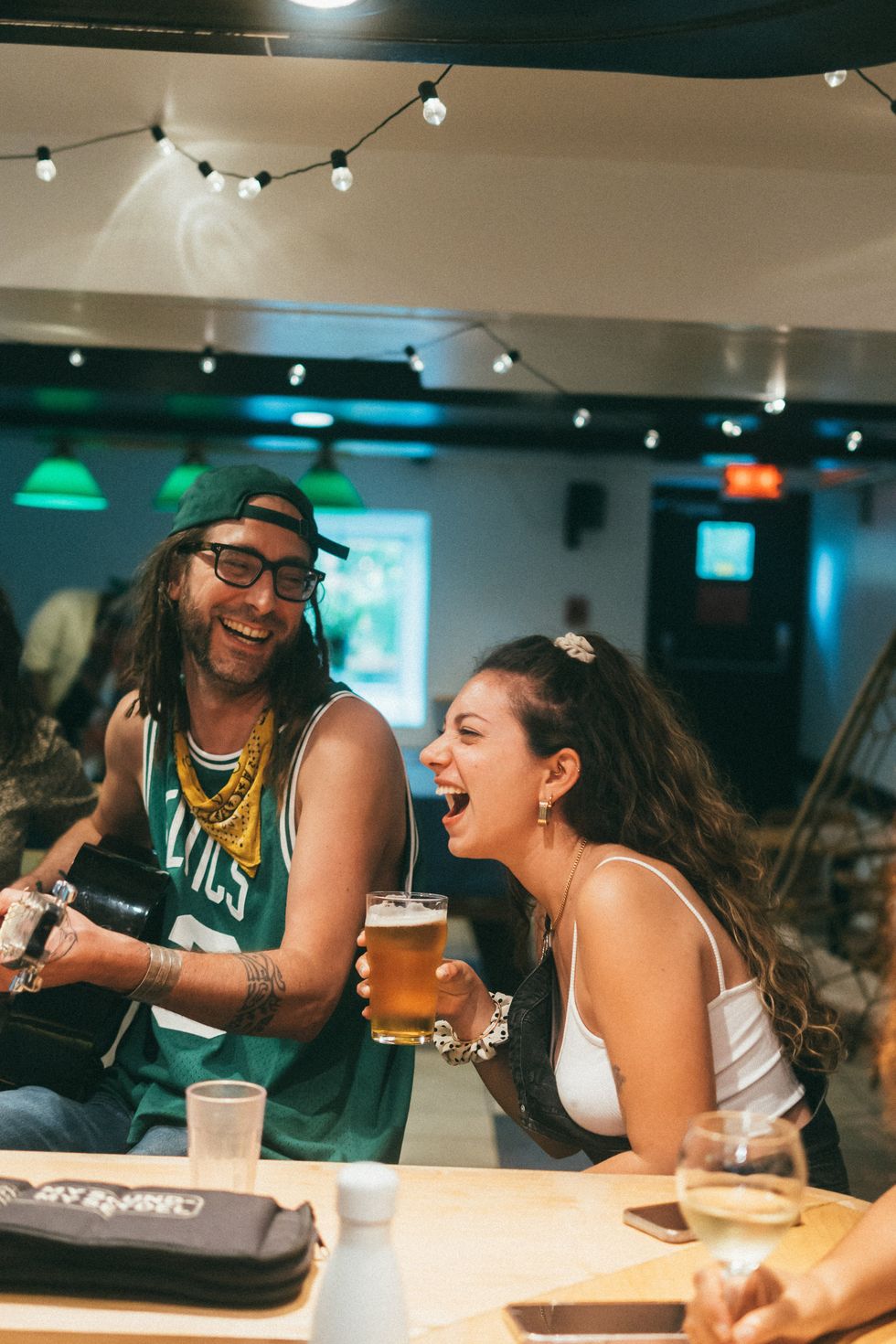 A man in a basketball singlet plays a guitar and smiles at a laughing woman in a white camisole who is drinking a beer. 