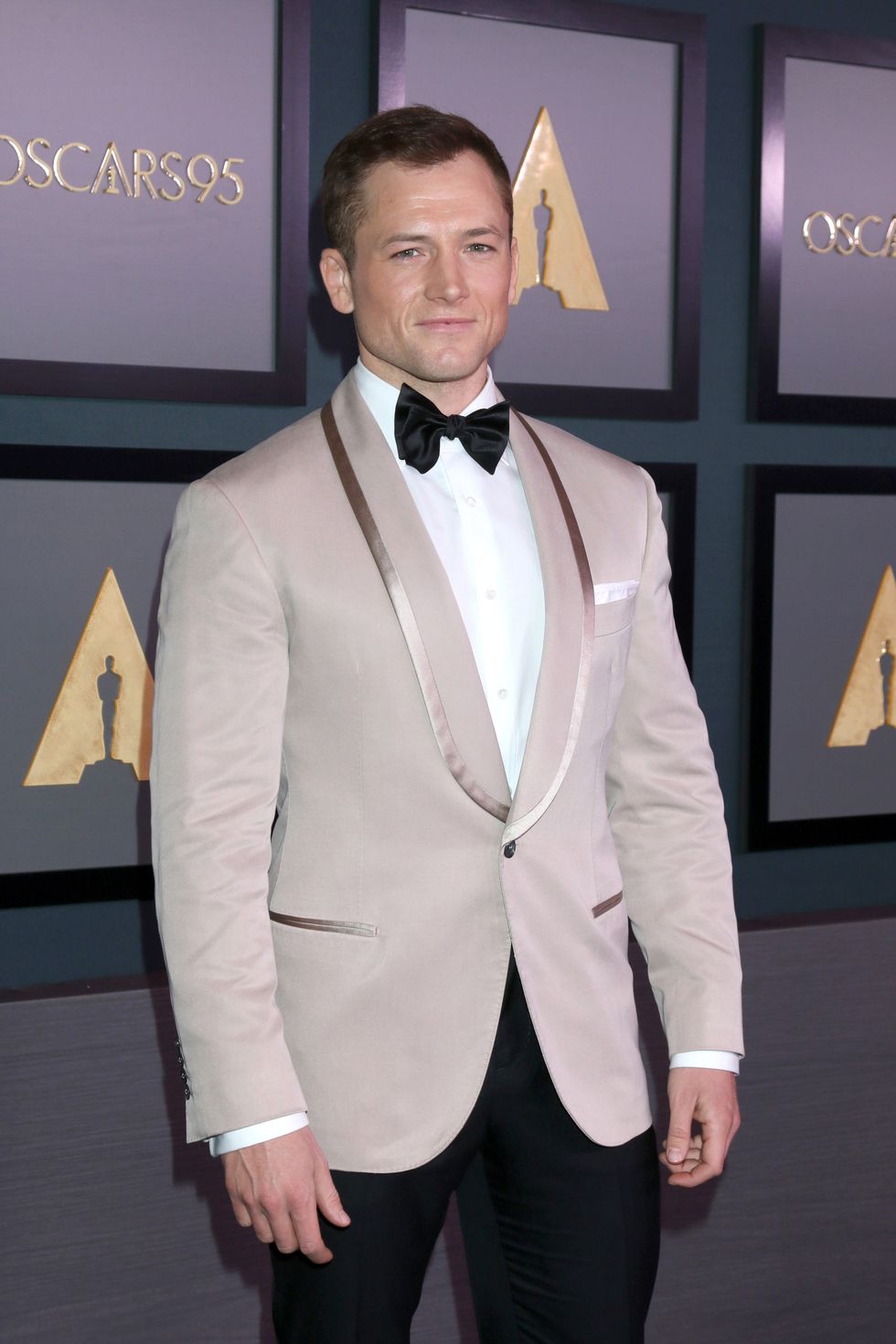 A man in a beige suit jacket, white dress shirt and black bowtie posing for a photo.