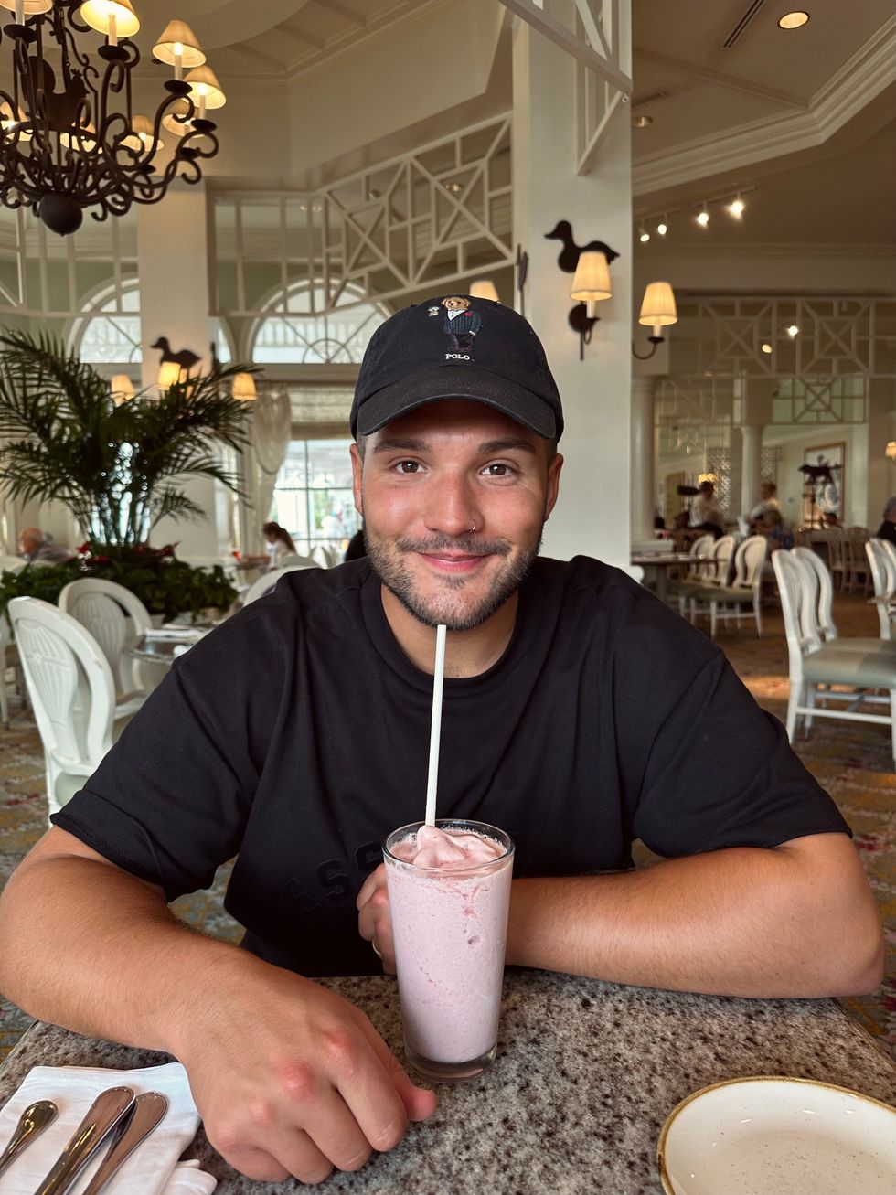A man in a black t-shirt and Polo cap smiling at the camera while drinking a pink milkshake through a straw at the Grand Floridian Cafe, with the resort's airy white dining room visible in the background.