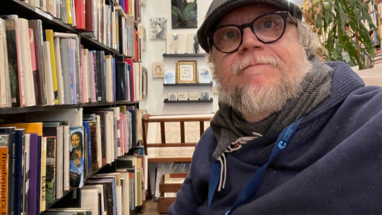 A man in a cap and glasses taking a selfie next to a bookshelf filled with books.