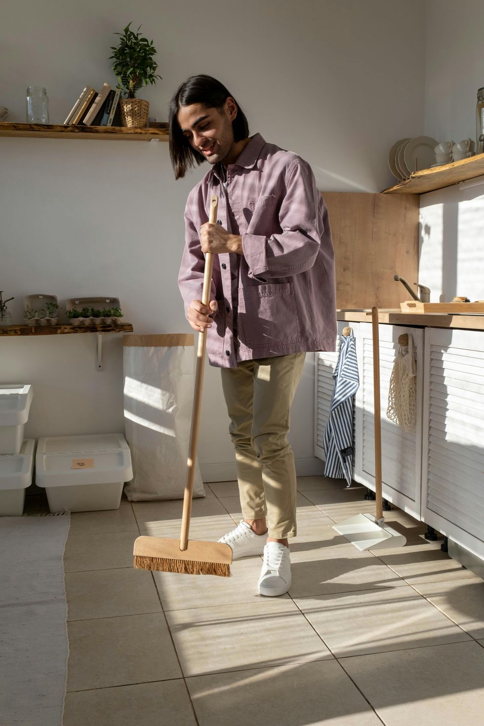 A man in a purple jacket, tan pants and white sneakers sweeps the tiled floor of a kitchen with white cabinets and wood countertops. 
