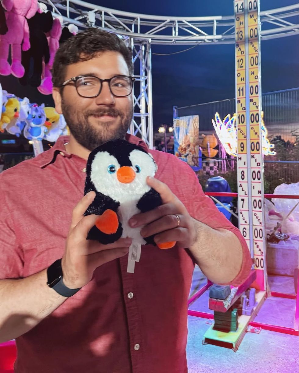 A man in a red button-down shirt holds a plushie penguin in front of a strength tester sideshow attraction. 