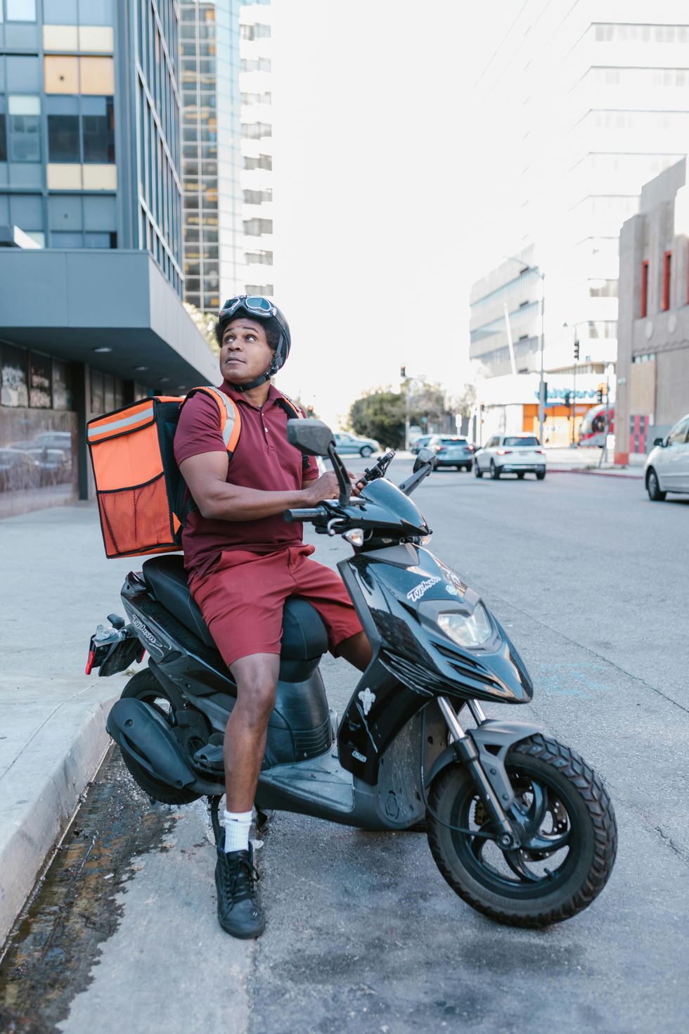 A man in maroon shorts and tank and wearing a delivery backpack sits astride a scooter and checks his phone.