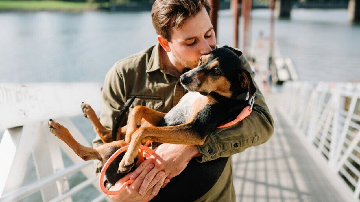 A man on a bridge holding a dog in his arms kisses the dog on the side of the face.